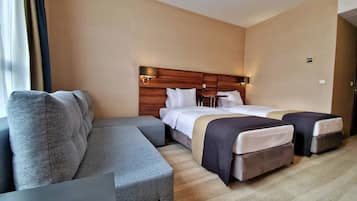 Une chambre d’hôtel avec un canapé, deux lits, une tête de lit en bois et une lampe fixée au mur.