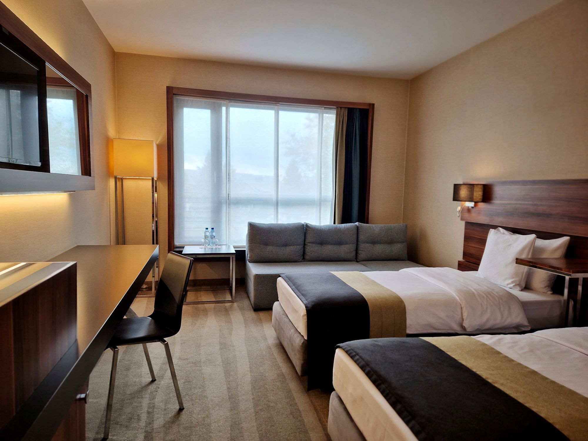 Ein Hotelzimmer mit Schreibtisch, zwei Betten, einer Couch, einem Fenster mit Vorhängen und einer Lampe.