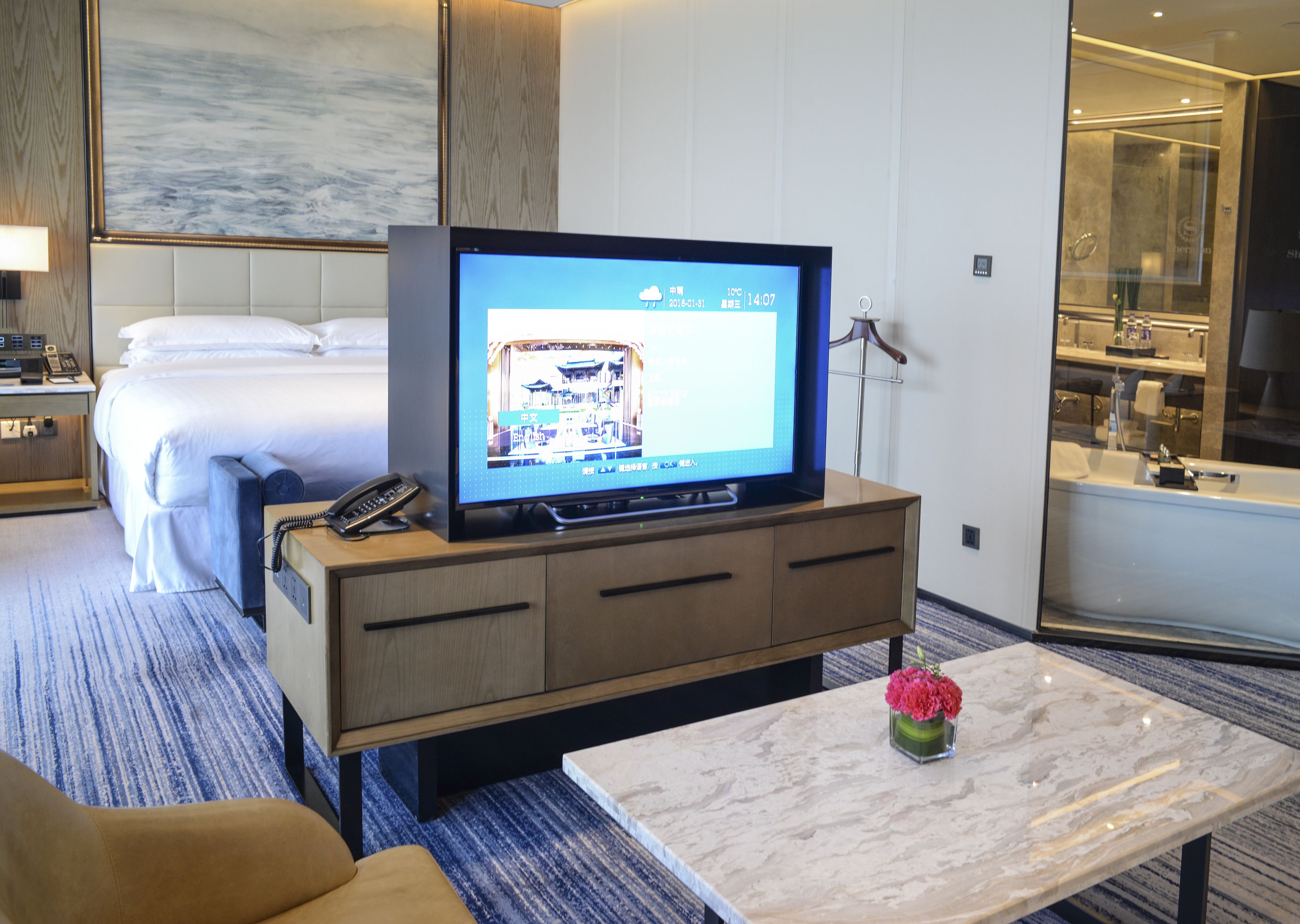 Una habitación de hotel con una cama, un televisor sobre un mueble de madera, un baño con separador de vidrio y una mesa de centro de mármol con un jarrón de flores.