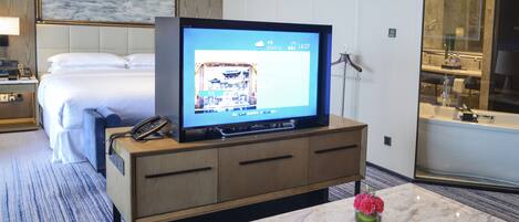 Una habitación de hotel con una cama, un televisor sobre un mueble de madera, un baño con separador de vidrio y una mesa de centro de mármol con un jarrón de flores.