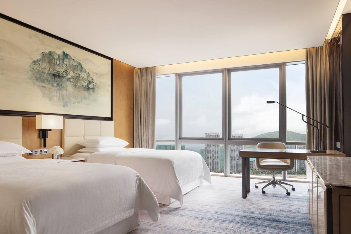 Ein Hotelzimmer mit zwei Betten, einem Schreibtisch, einer Lampe und einem großen Fenster mit Blick auf die Stadt.