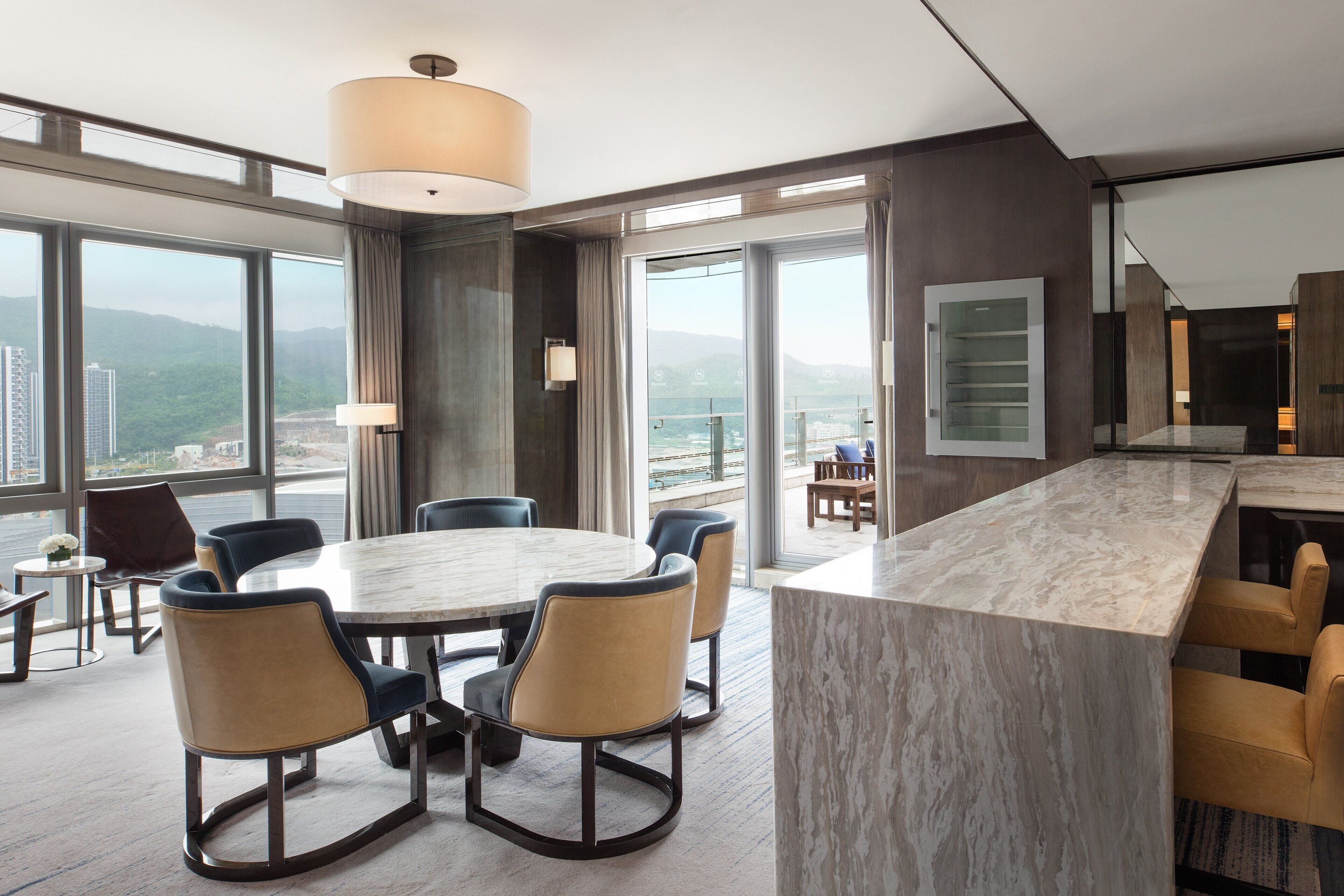A modern hotel room with a large dining table, comfortable seating, a large window with a city view, and a spacious balcony.