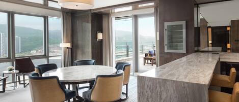 A modern hotel room with a large dining table, comfortable seating, a large window with a city view, and a spacious balcony.