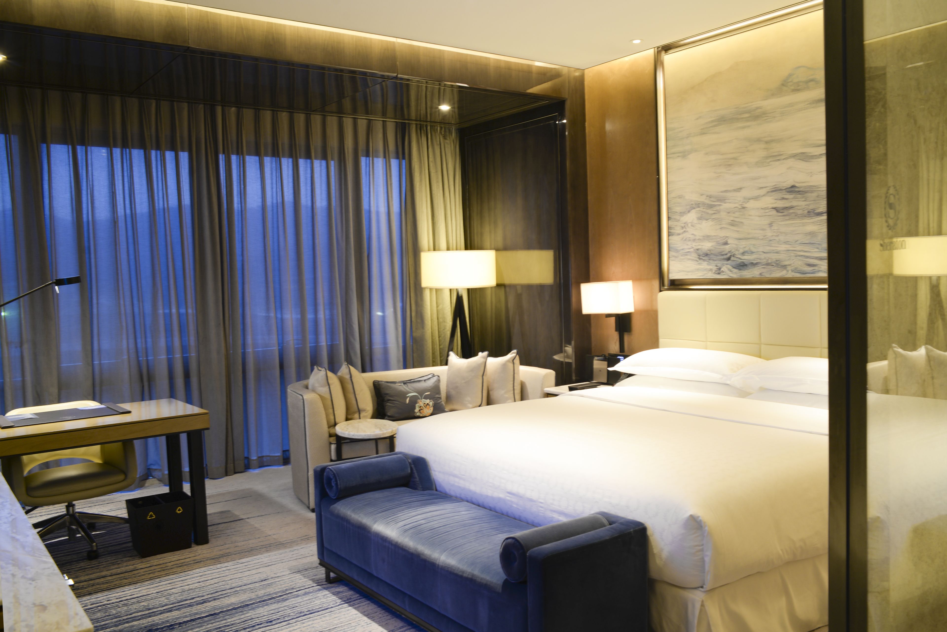 Una habitación de hotel moderna con una cama grande, un escritorio, un sofá y un ventanal con cortinas.