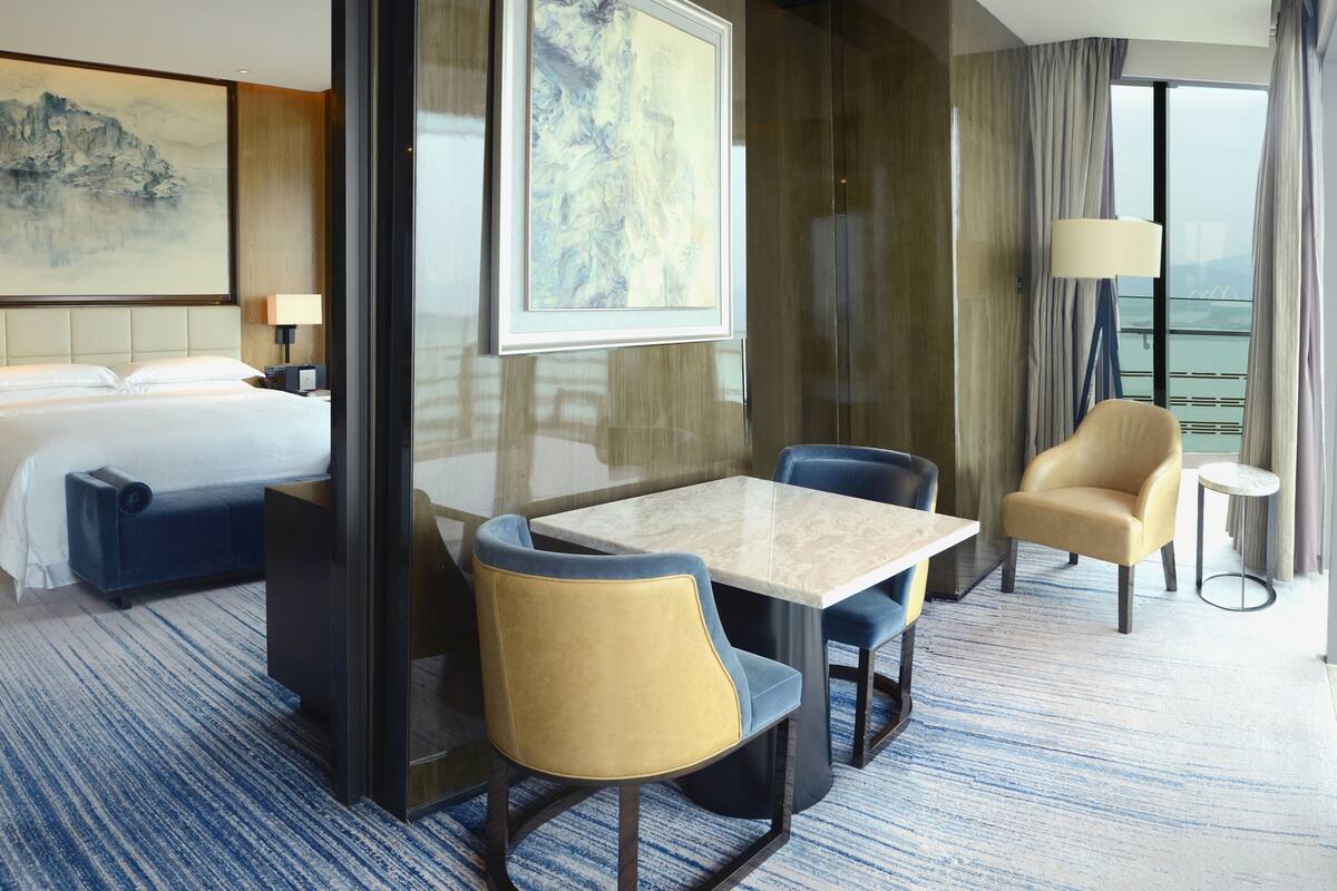 Ein Hotelzimmer mit einem Bett, einem Schreibtisch, zwei Stühlen, einem kleinen Tisch und einem großen Fenster mit Vorhängen.