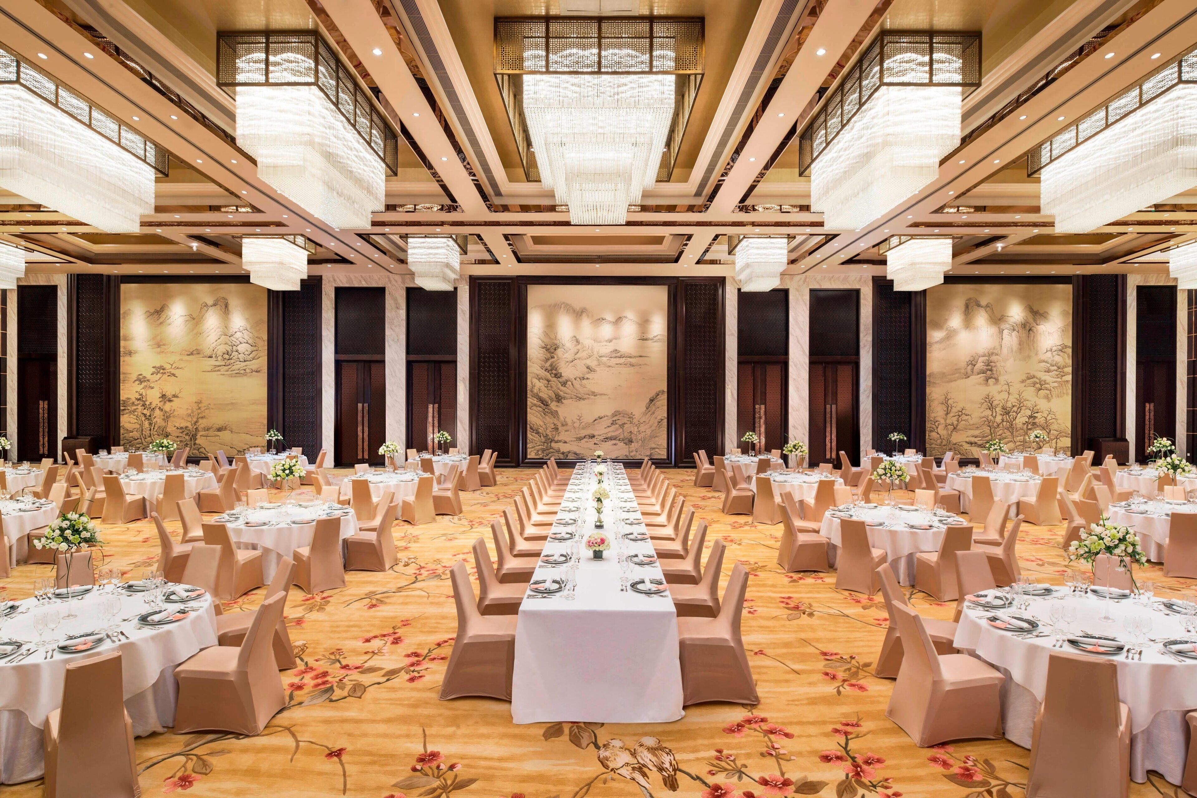 A banquet hall with round tables, beige chairs, and a long table with floral centerpieces.
