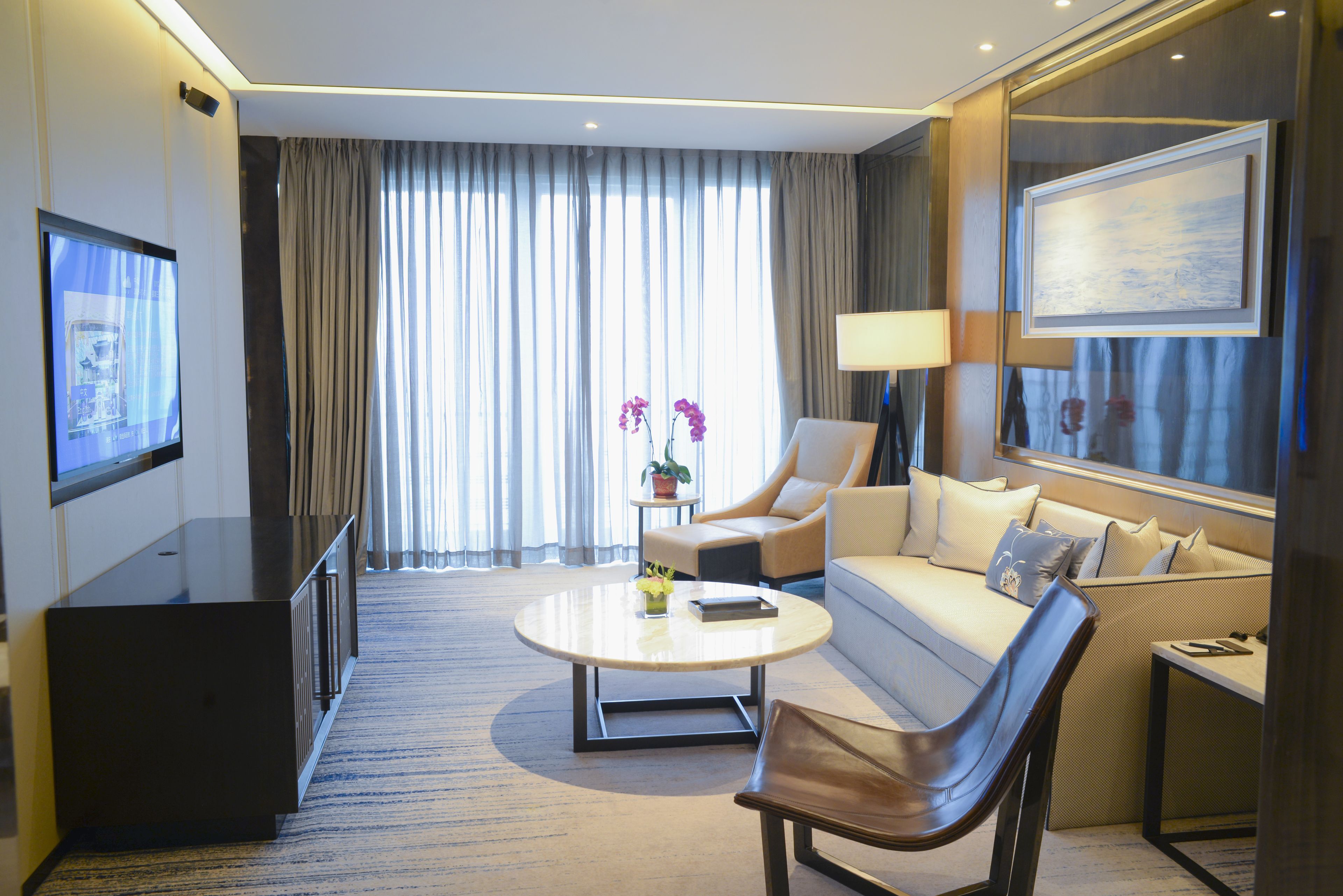 A modern hotel room with a flat-screen TV, a sofa, a chair, a coffee table, and a large window with curtains.