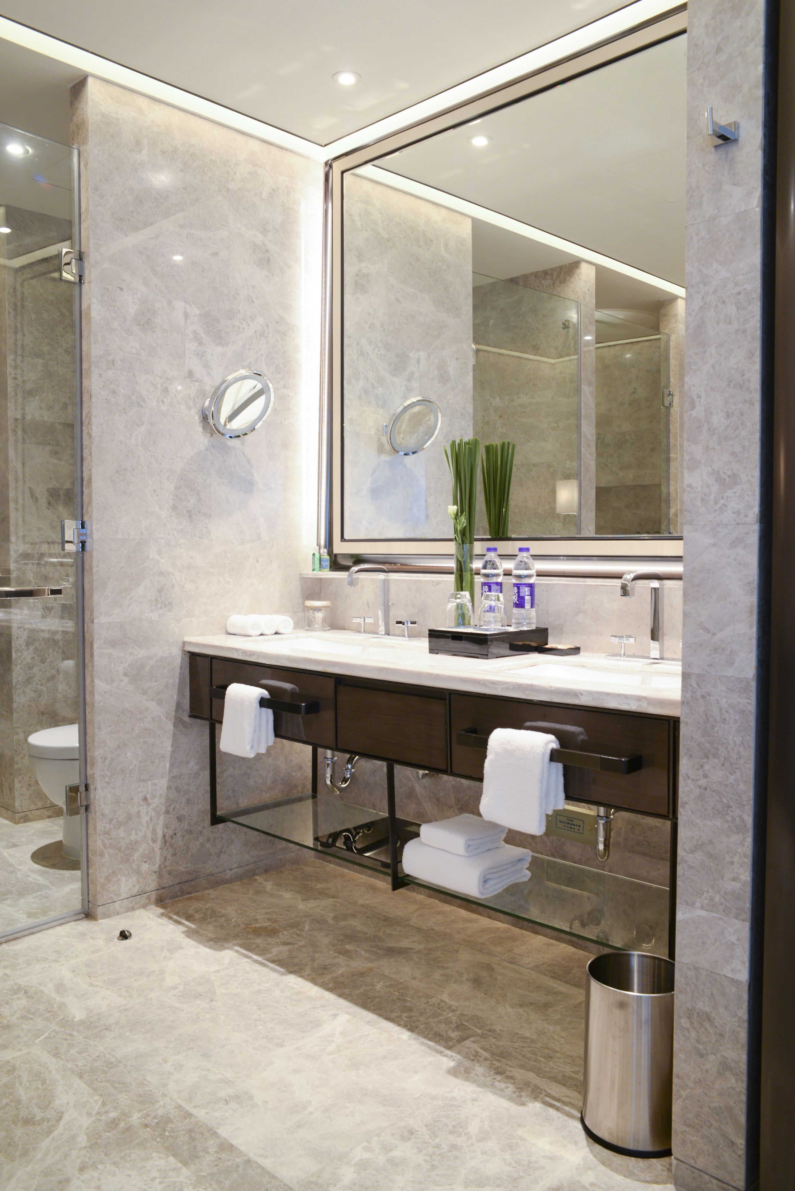 A modern bathroom with a large mirror, marble walls, a glass shower enclosure, and a stainless steel trash can.