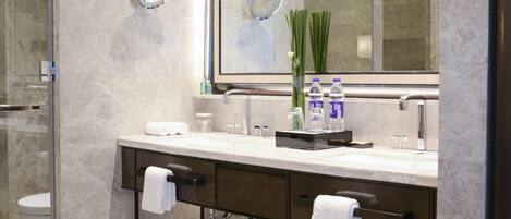 A modern bathroom with a large mirror, marble walls, a glass shower enclosure, and a stainless steel trash can.