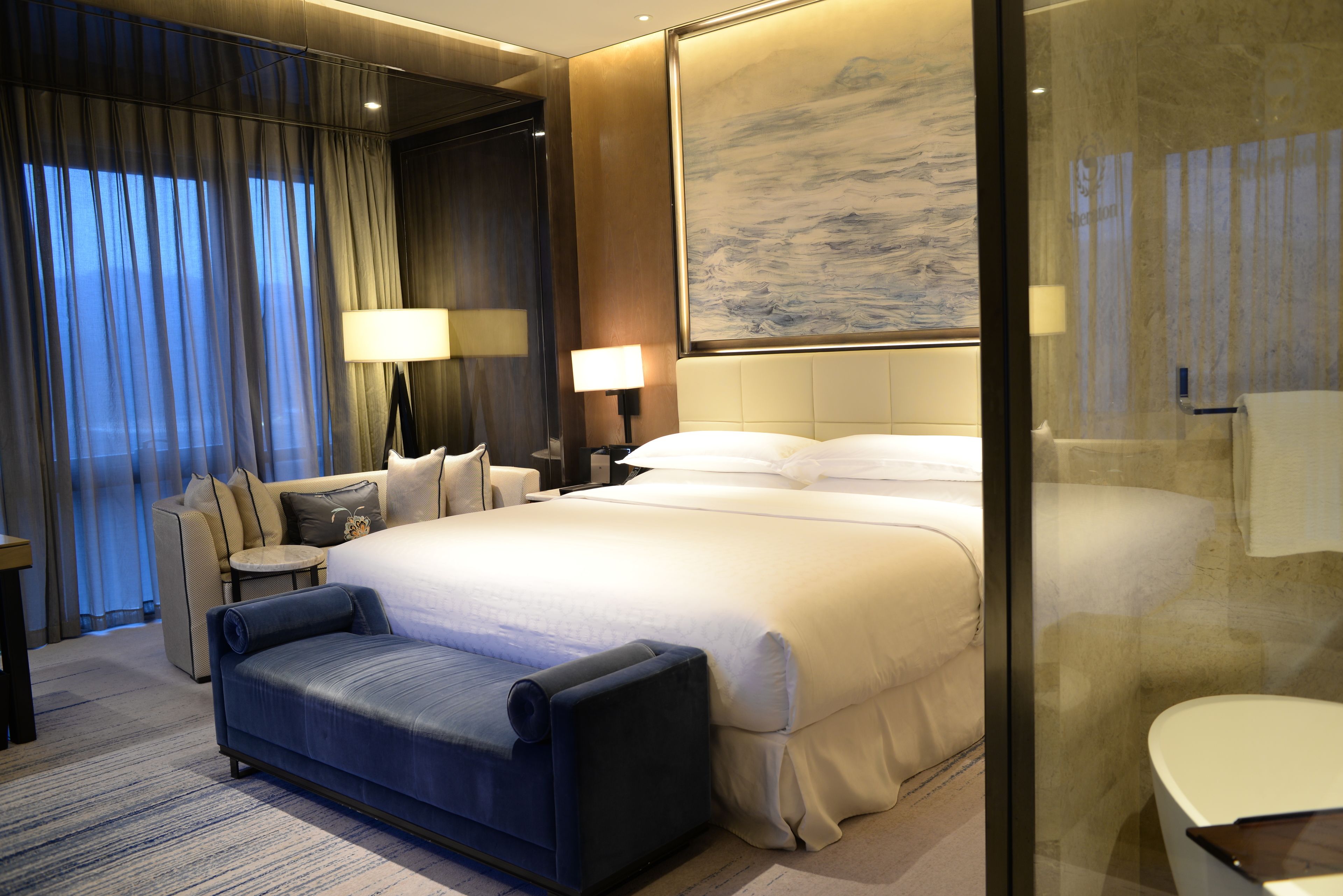 A modern hotel room with a large bed, a sofa, a desk, and a large window with curtains.