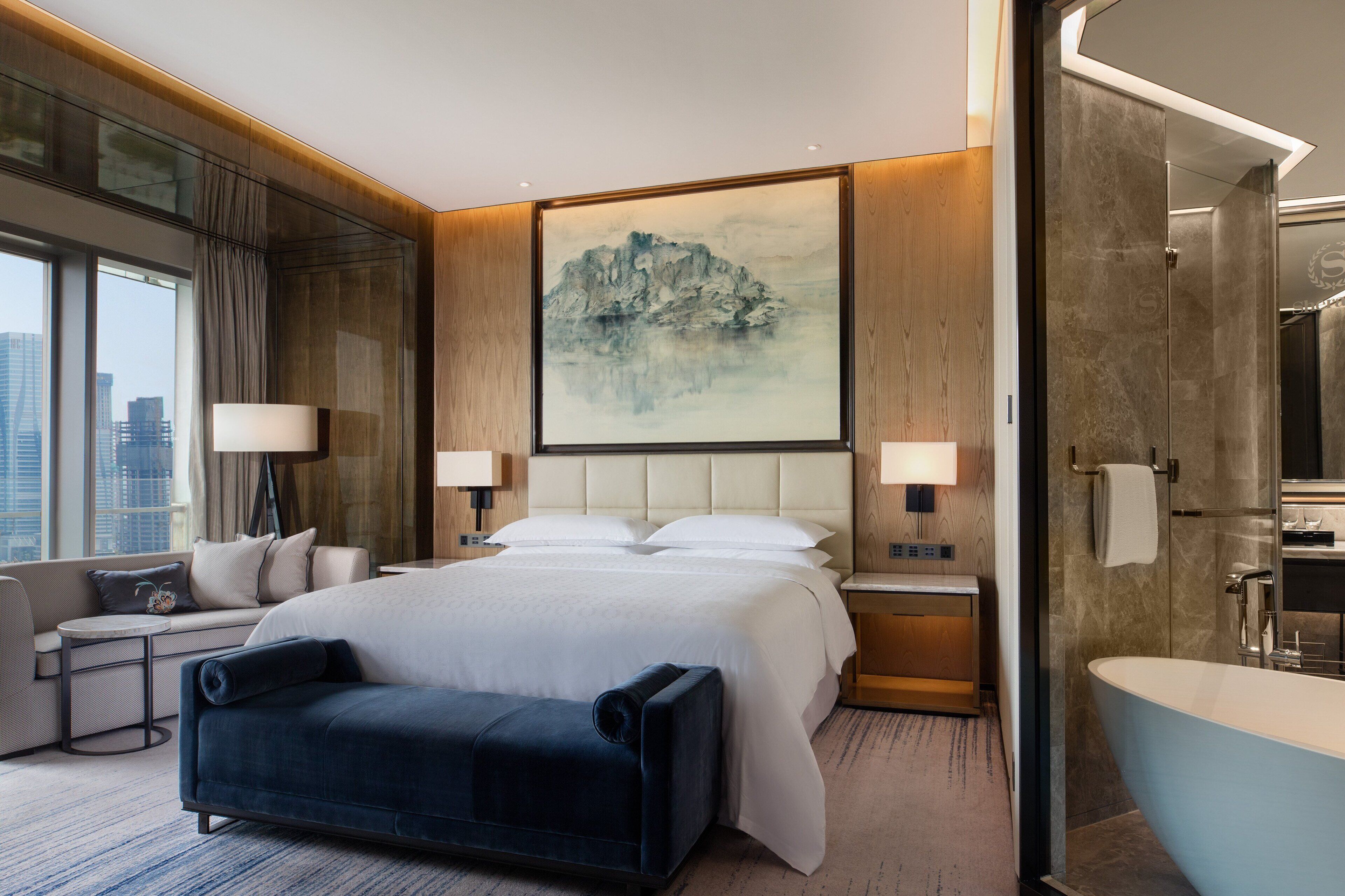 A modern hotel room with a large bed, a seating area, a bathroom with a bathtub, and a city view.