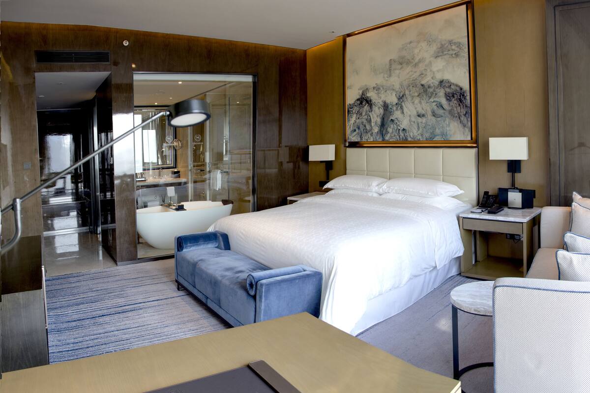 Ein modernes Hotelzimmer mit einem großen Bett, einer Sitzecke mit Sessel und Hocker, einem Schreibtisch und einem Badezimmer mit Badewanne.