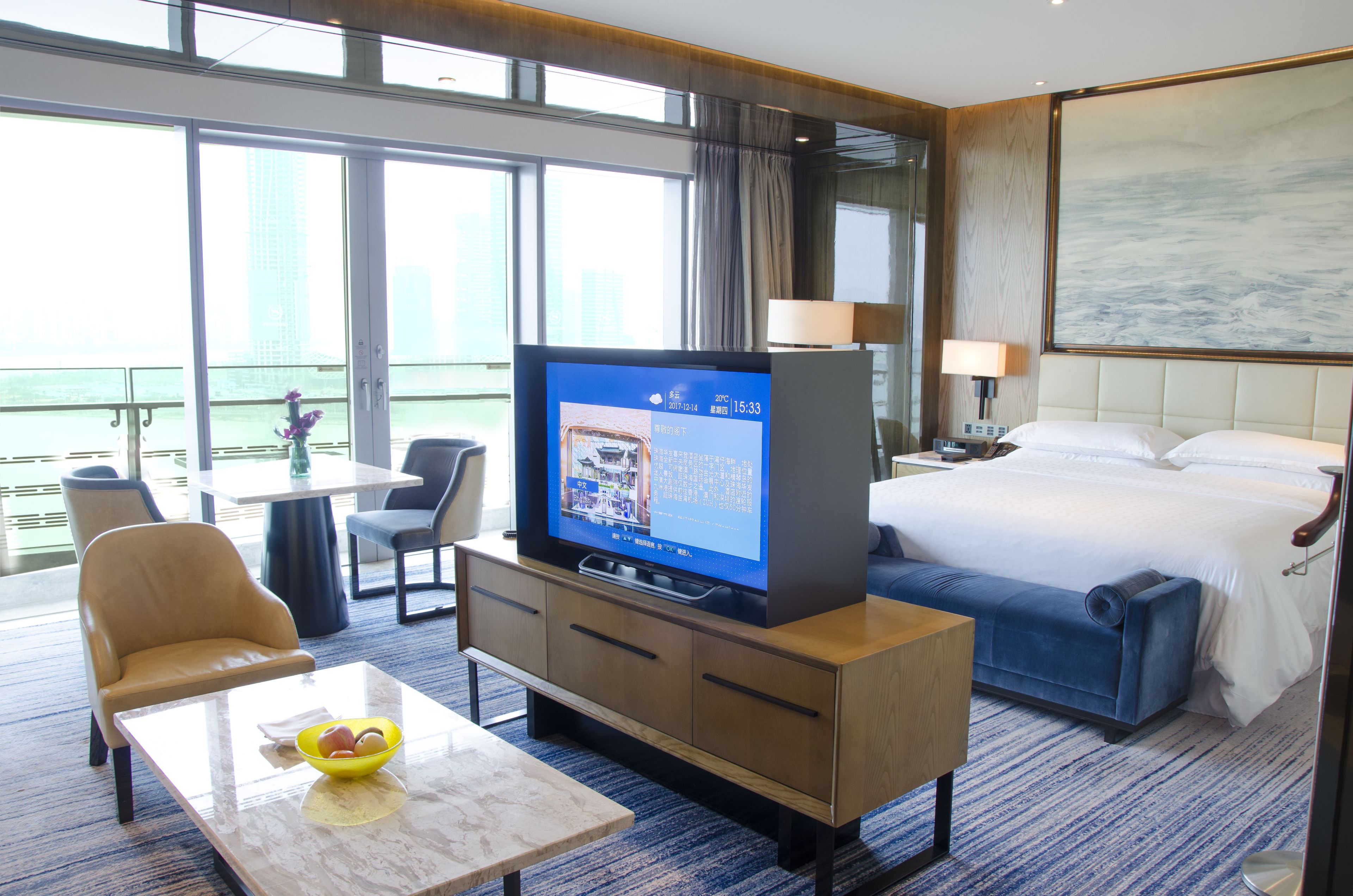 A hotel room with a bed, a TV on a wooden stand, a desk with a chair, a small table with a bowl, and a view of the city through large windows.