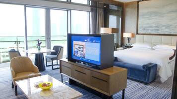 A hotel room with a bed, a TV on a wooden stand, a desk with a chair, a small table with a bowl, and a view of the city through large windows.
