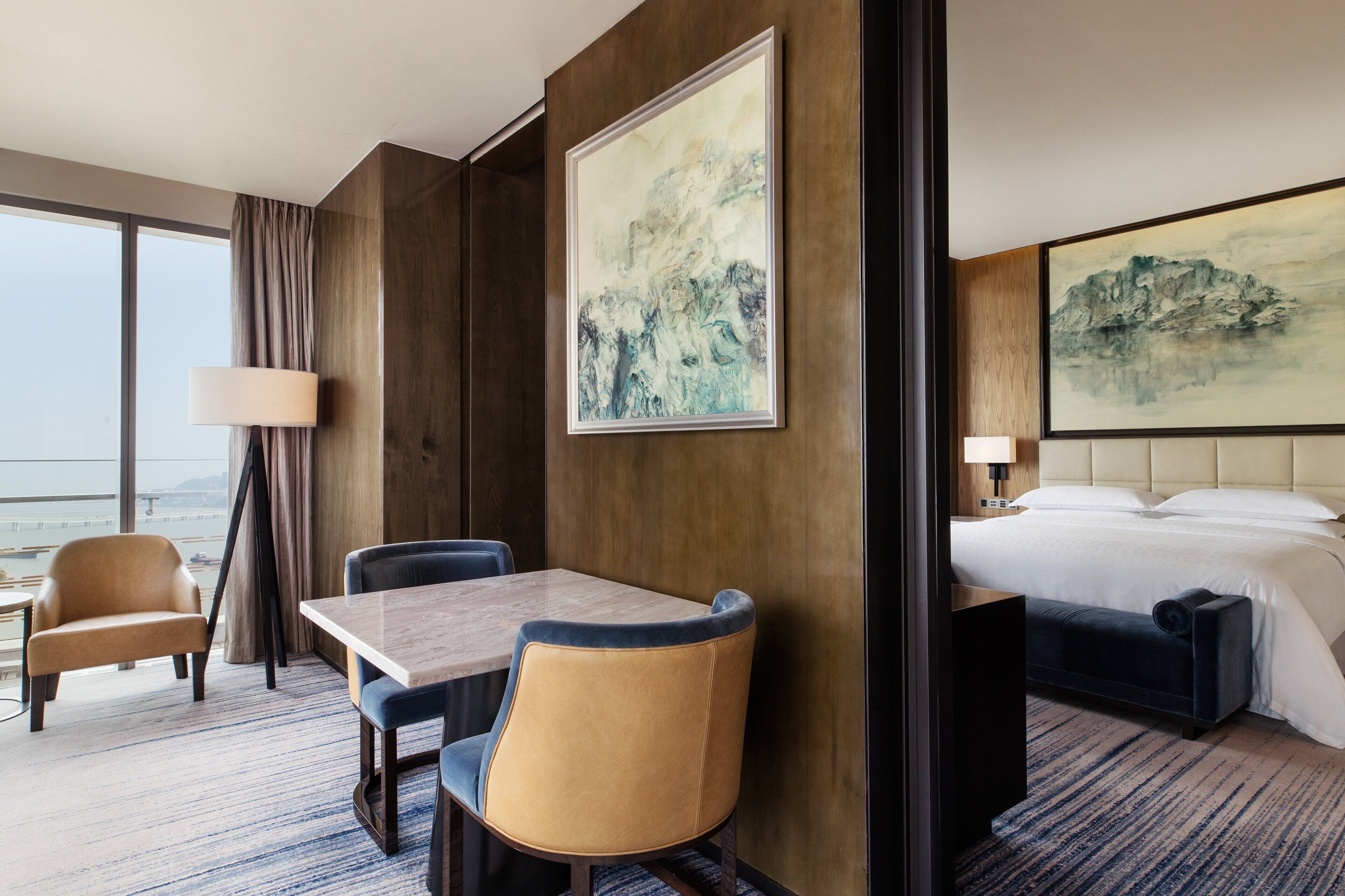 A hotel room with a large window, a bed, a desk, two chairs, and two paintings on the walls.