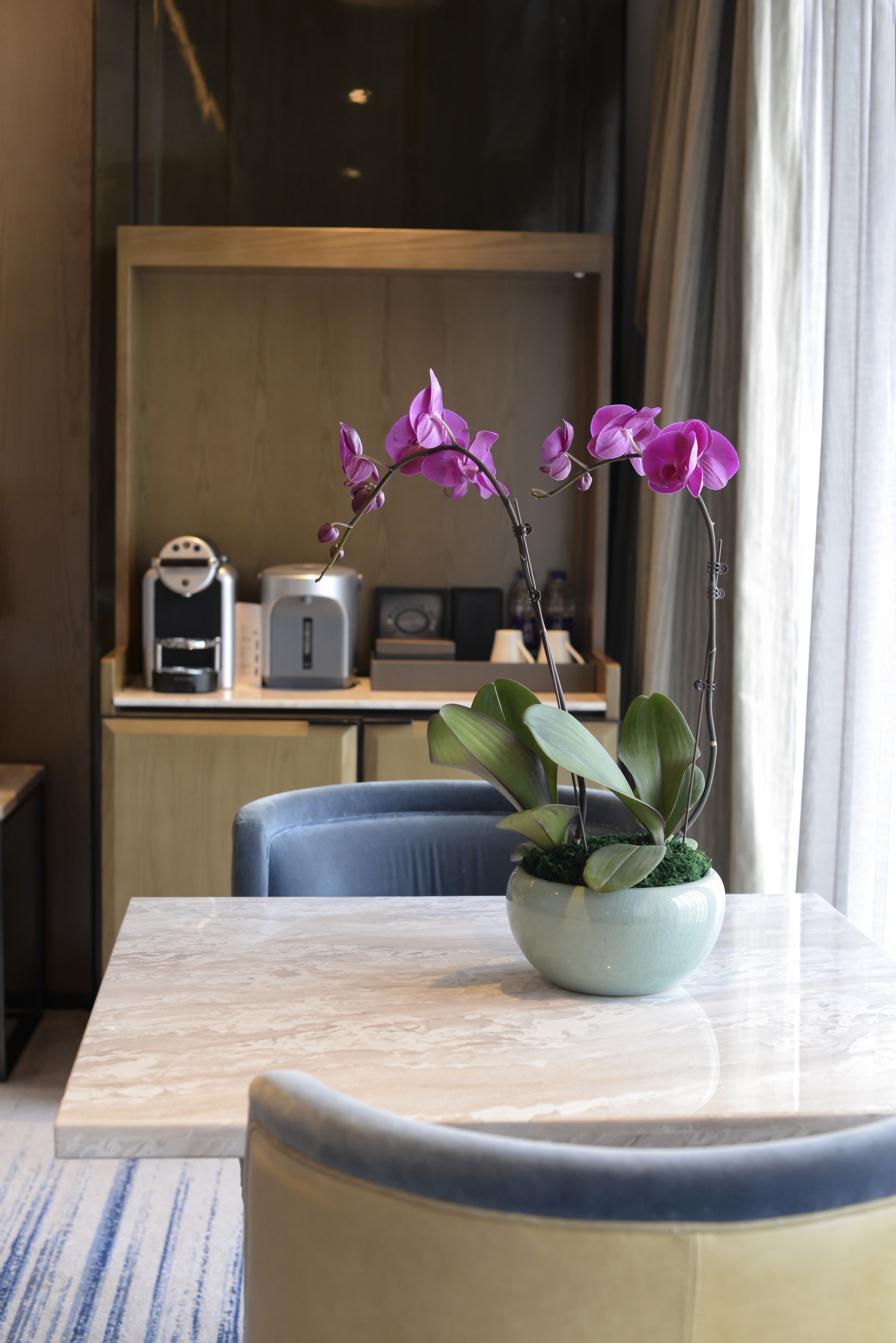 A hotel room with a marble table, a potted orchid, a blue chair, and a kitchenette in the background.