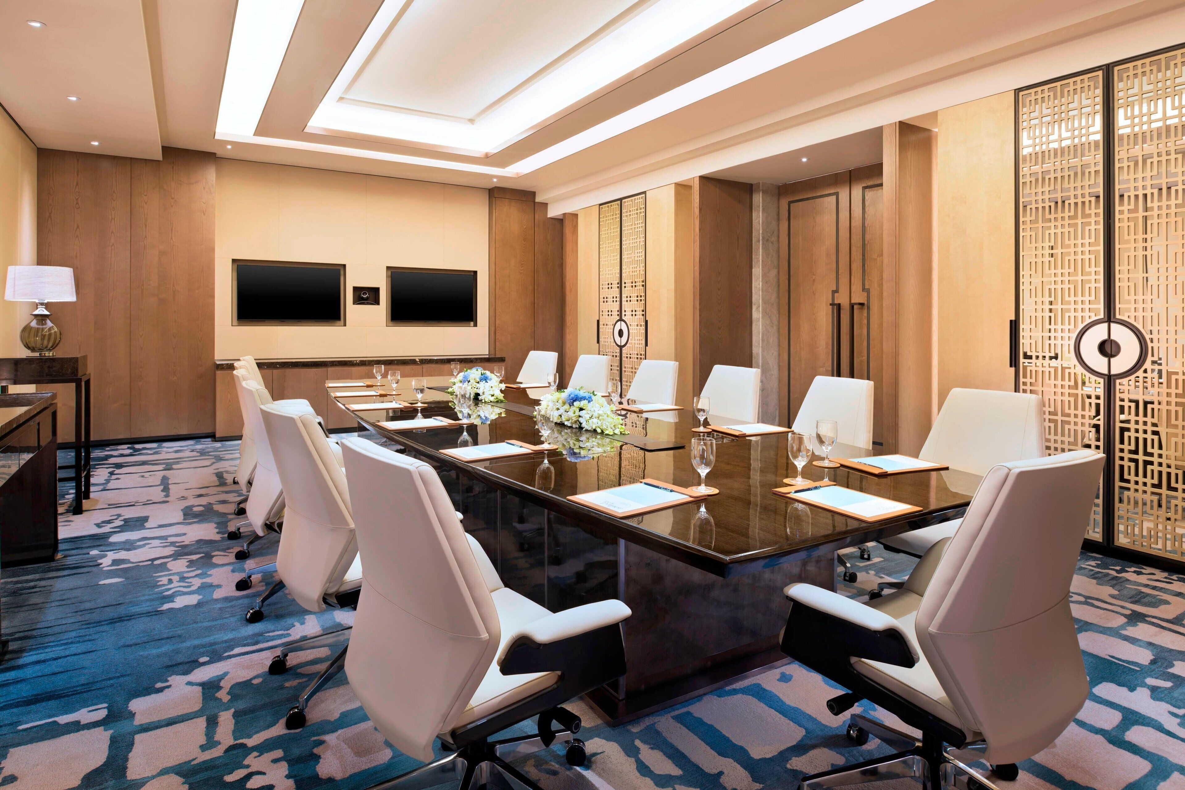 A conference room with a long table, white chairs, a TV, and a decorative screen.