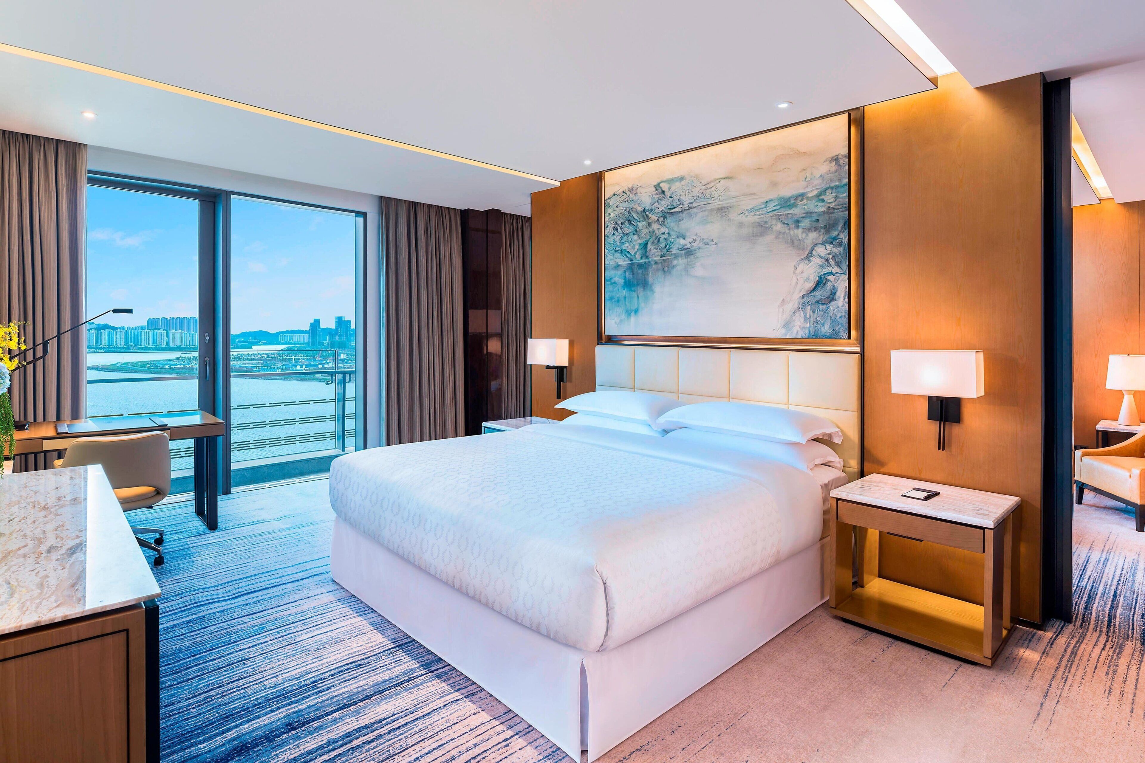 A modern hotel room with a large bed, a desk, and a view of the cityscape.