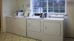 A laundry area with four white washers and dryers, a window with a view of greenery, and an attention sign on the wall. - Lighthouse Inn (Crescent City)