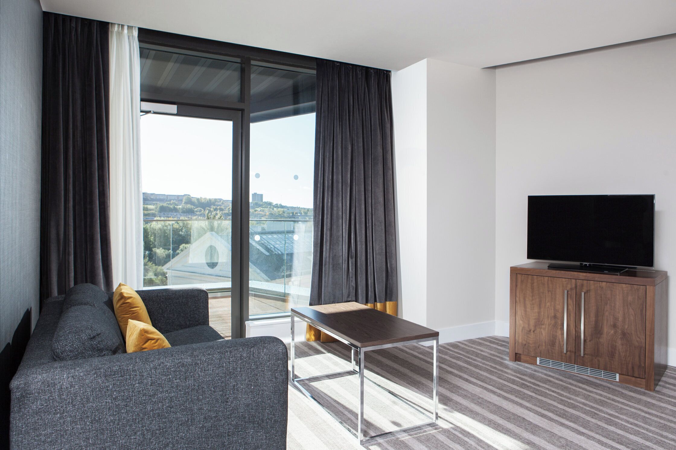 A modern hotel room with a sofa, a small table, a television, and a view of the cityscape.
