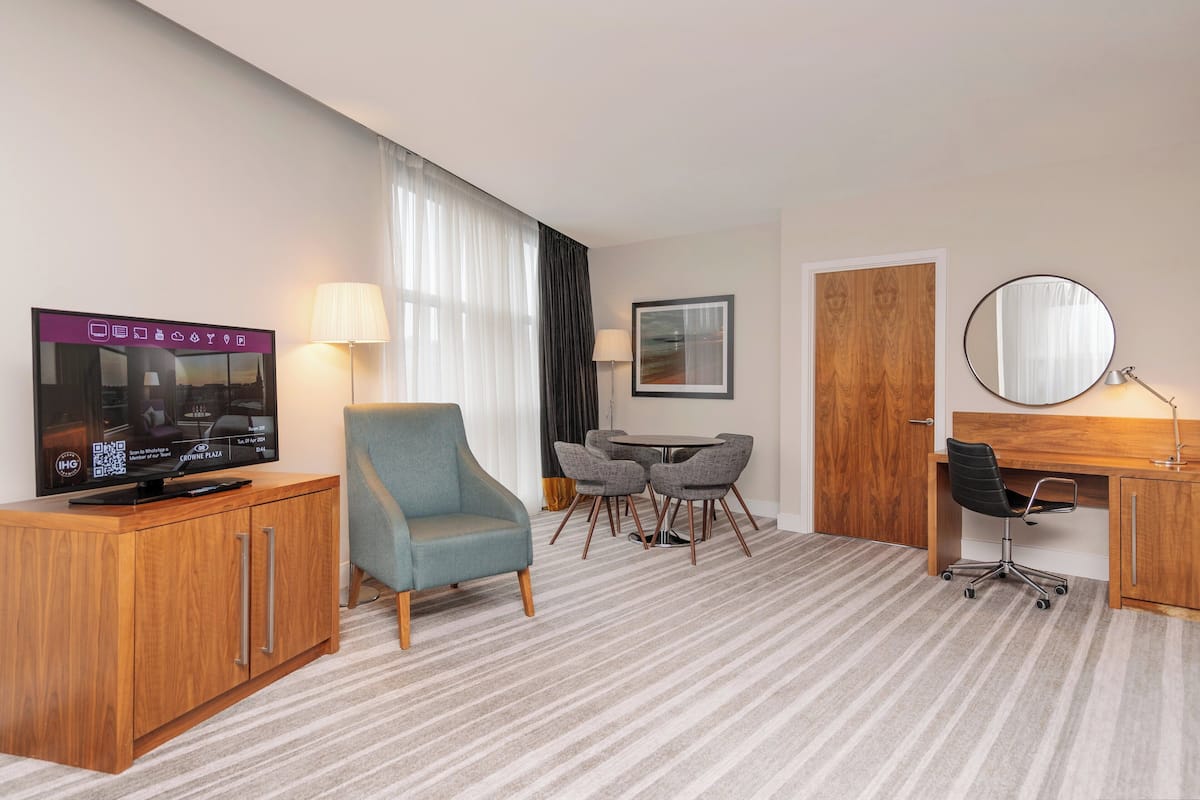 A hotel room with a wooden cabinet, a television, a blue armchair, a dining table with chairs, a desk with a chair, and a round mirror.