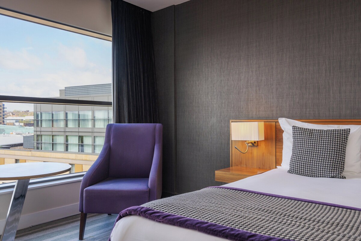 Een hotelkamer met een bed, een paarse fauteuil, een ronde tafel en een uitzicht op gebouwen door grote ramen.