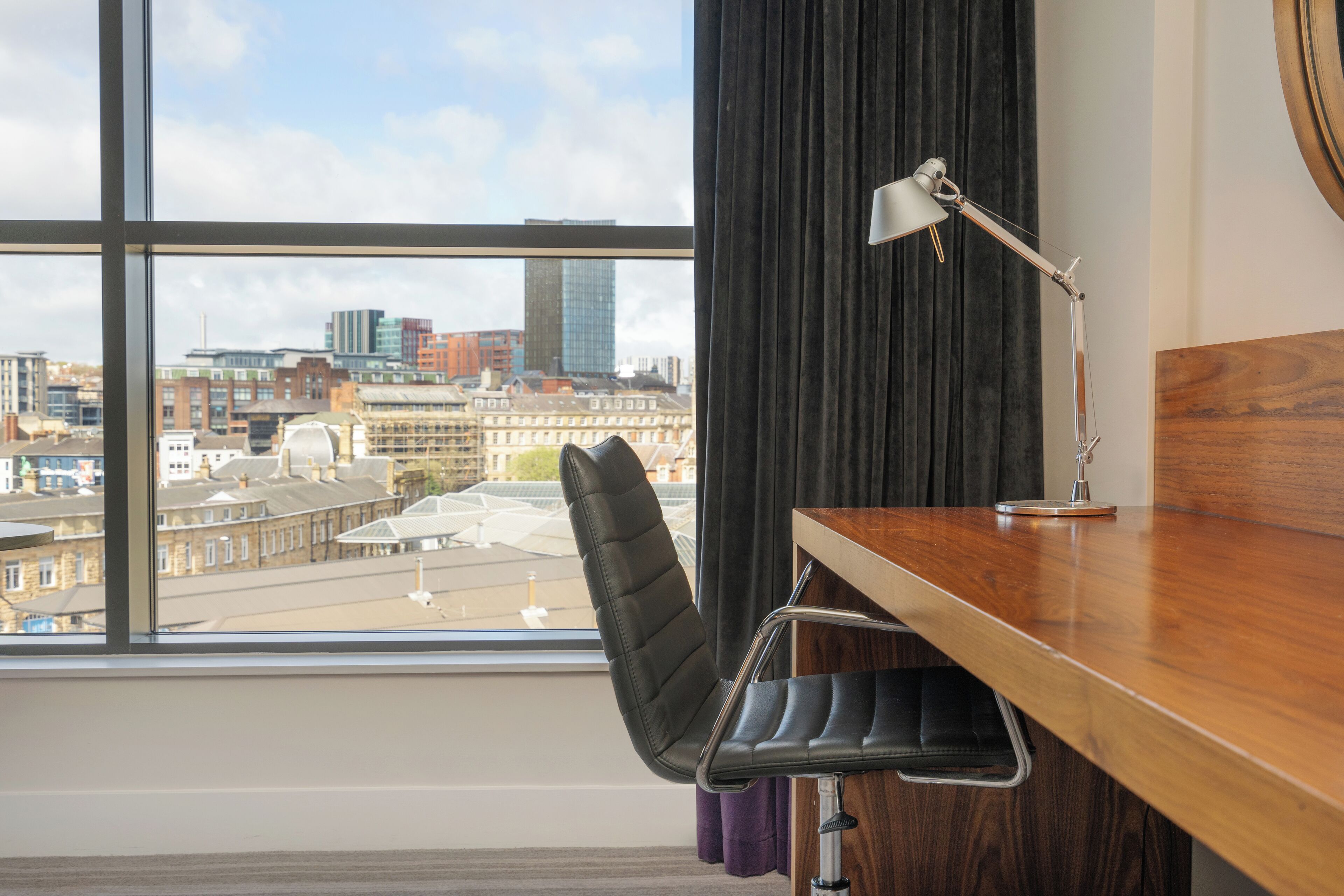 A modern office chair with a high backrest and metal frame, positioned in front of a wooden desk with a desk lamp. A large window provides a view of a cityscape with buildings and a clear sky.