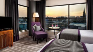A hotel room with a large window, a flat-screen TV, a purple armchair, a small round table with a lamp, and a bed with patterned bedding.