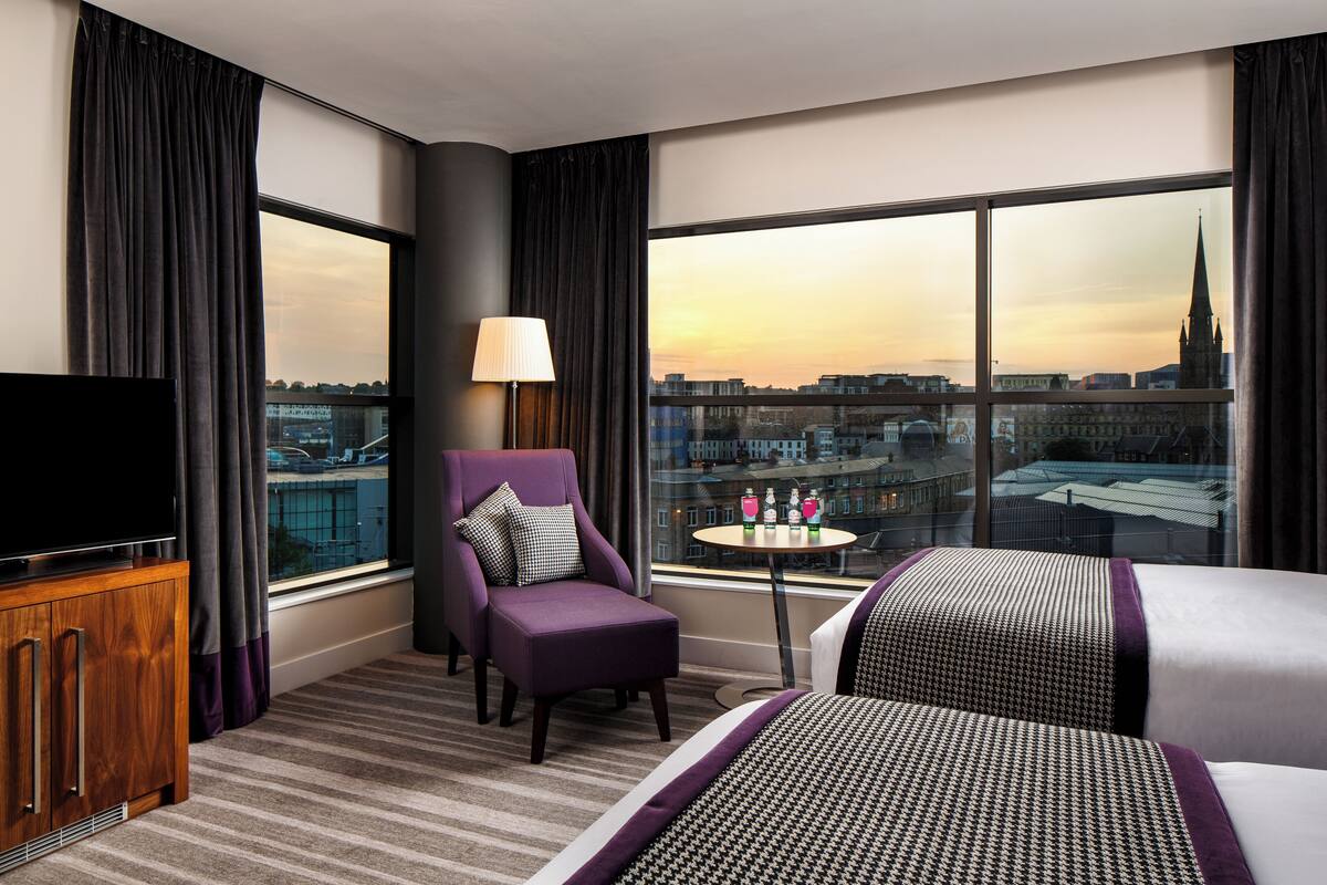 A hotel room with a large window, a flat-screen TV, a purple armchair, a small round table with a lamp, and a bed with patterned bedding.