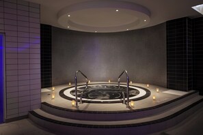 Een moderne spa met een rond bubbelbad, een trap die er naartoe leidt, en kaarsen die voor sfeerverlichting zorgen.