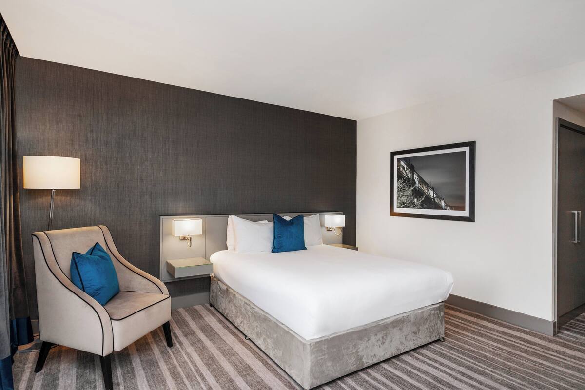 A hotel room with a large bed, a chair, a nightstand, a lamp, and a framed picture on the wall.