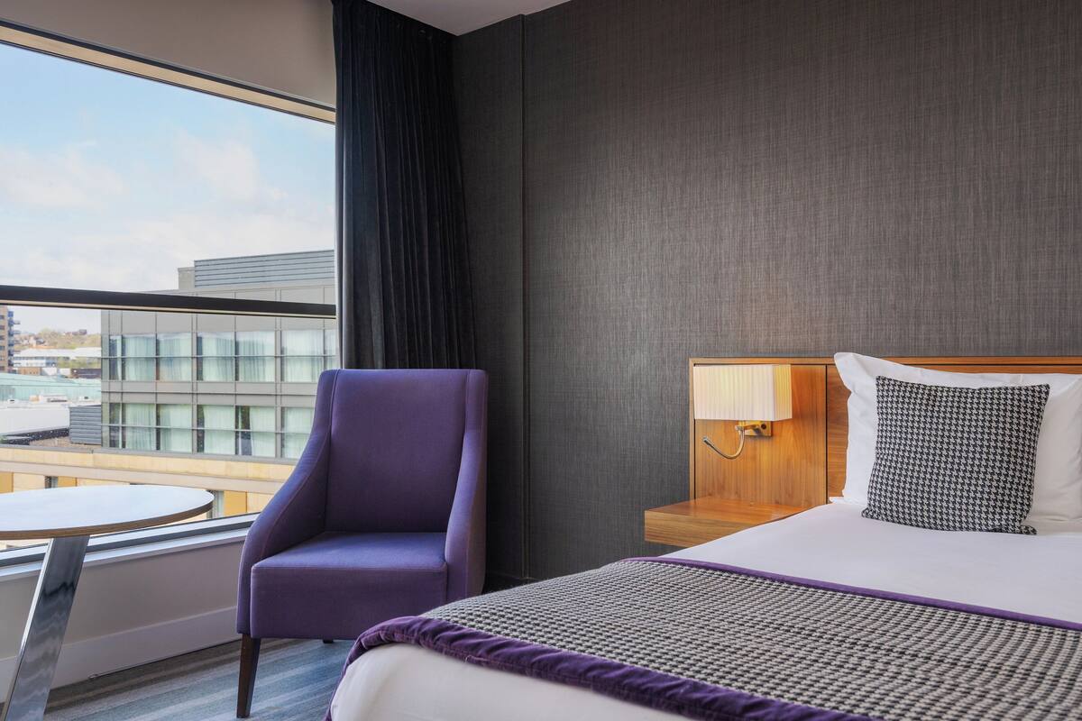 A hotel room with a bed, a purple armchair, a round table, and a view of buildings through large windows.