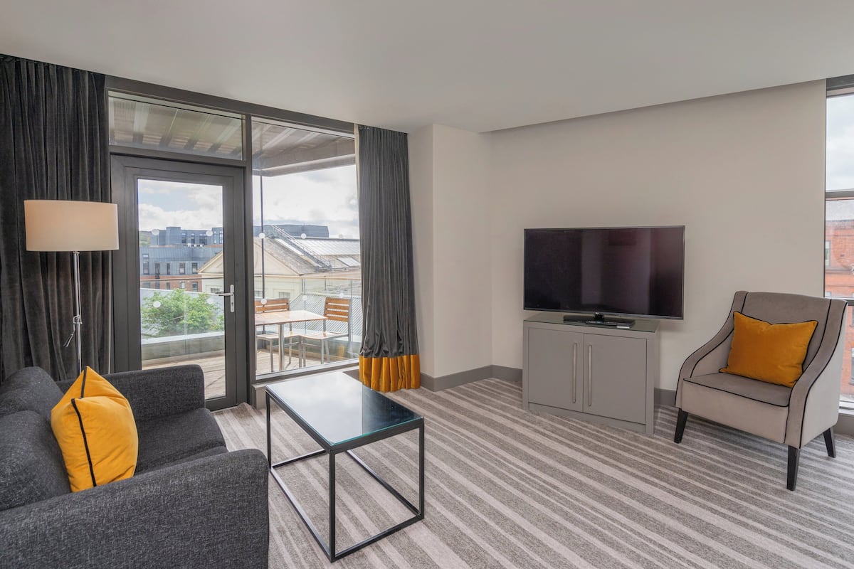 A modern hotel room with a sofa, a small table, a TV, and a view of the city through large windows.