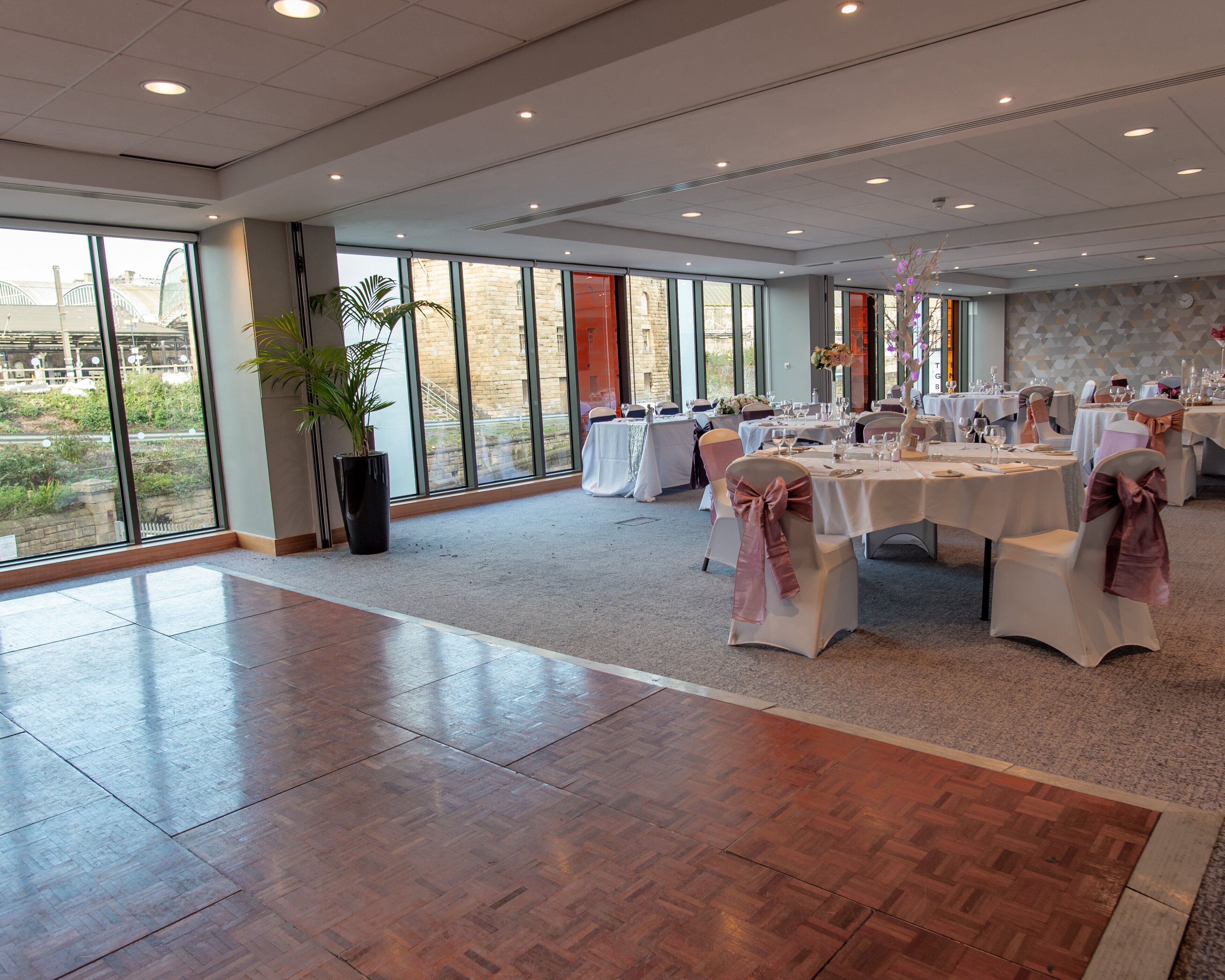 A spacious banquet hall with large windows, polished wooden floors, and tables set for a formal event.