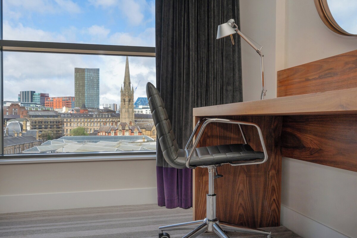 Een moderne hotelkamer met een groot raam en uitzicht over de stad, een houten bureau, een zwarte bureaustoel en een vloerlamp.