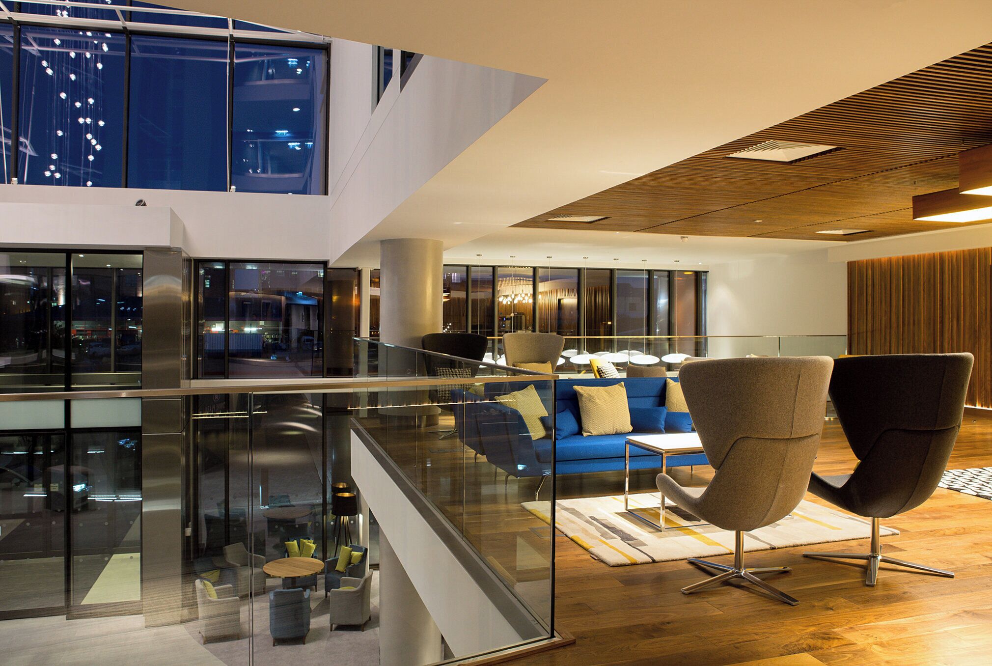 Een modern interieur met een glazen balustrade, een blauwe bank en een hoog plafond met houten balken.