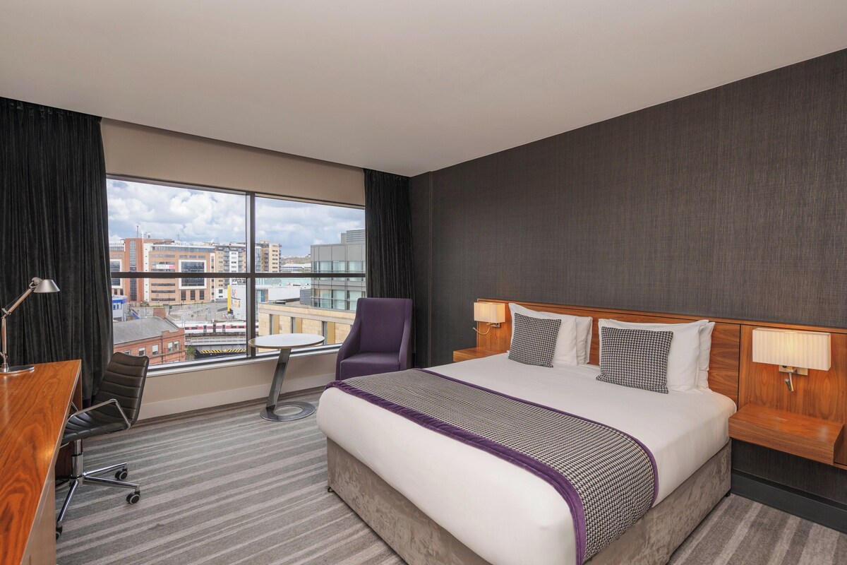 Een hotelkamer met een groot bed, een bureau, een stoel en uitzicht op de stad door het raam.