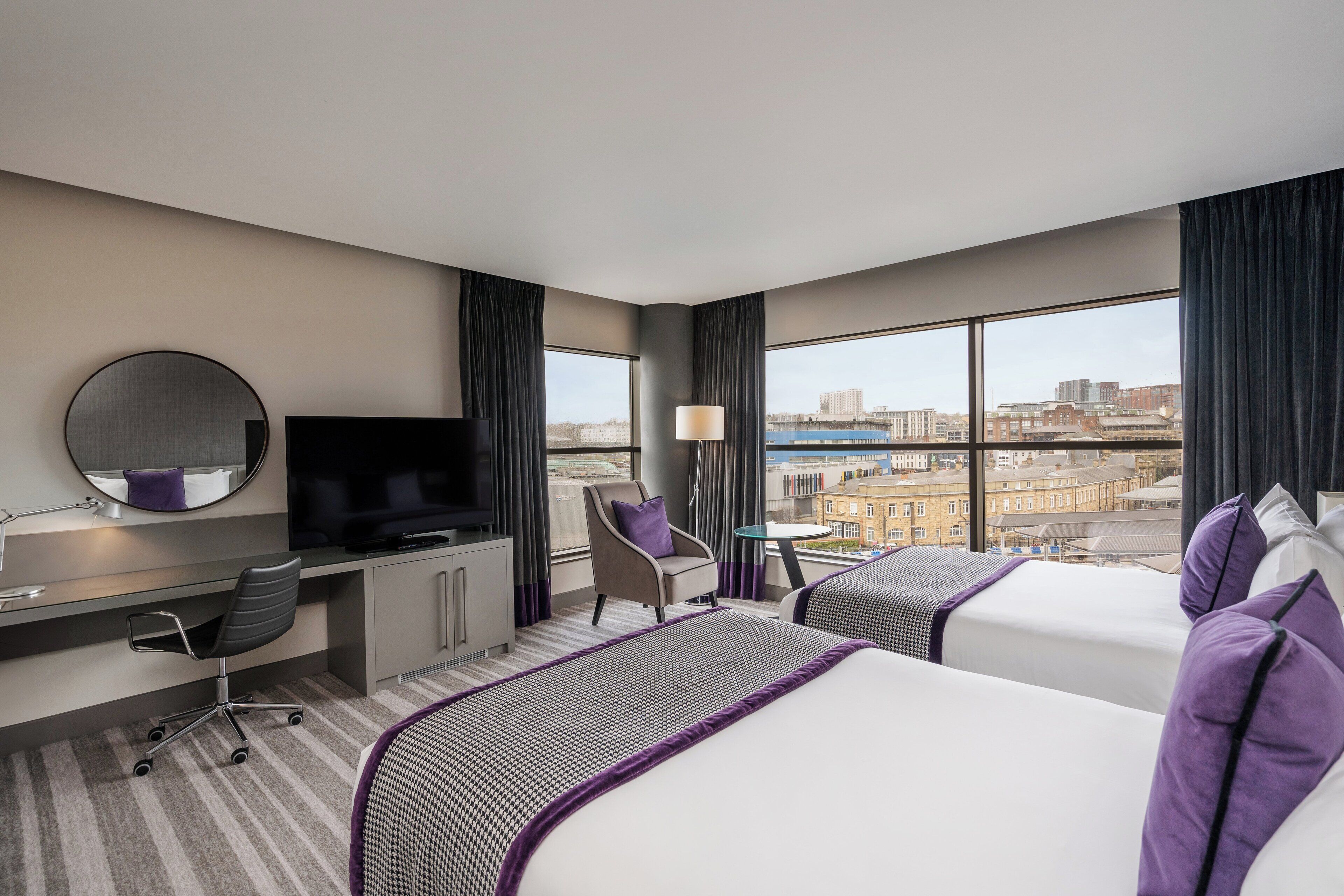 A hotel room with two beds, a desk, a chair, a TV, and large windows offering a city view.