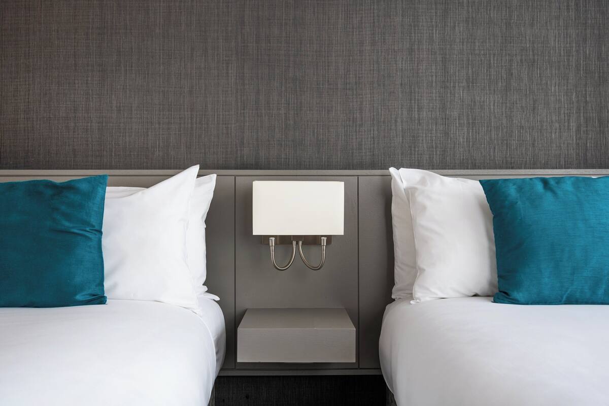 A hotel room with two beds, each with white bedding and teal pillows. There is a bedside table with a white lamp and a small drawer.