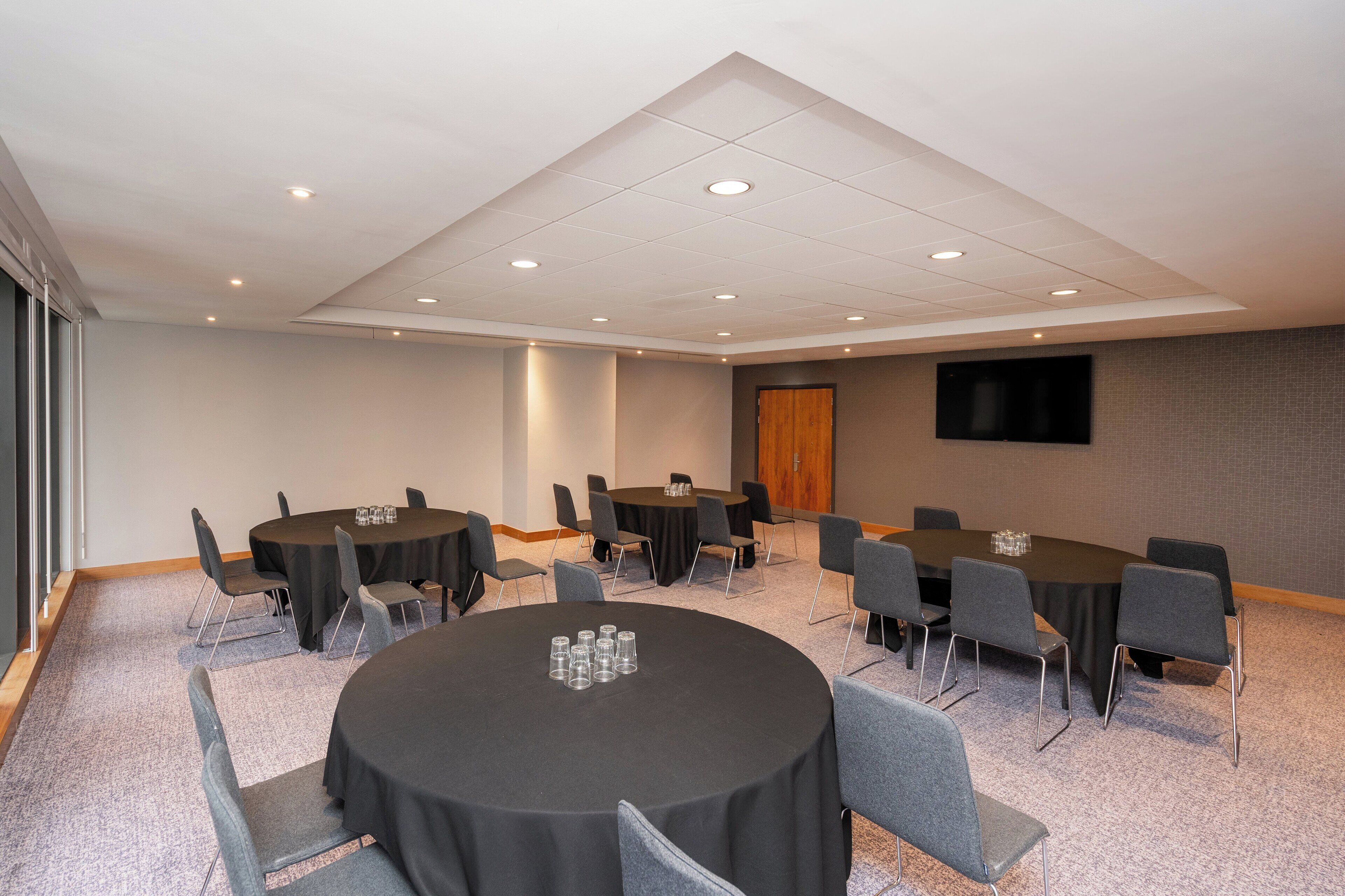 A conference room with multiple round tables, grey chairs, and a flat-screen TV on the wall.