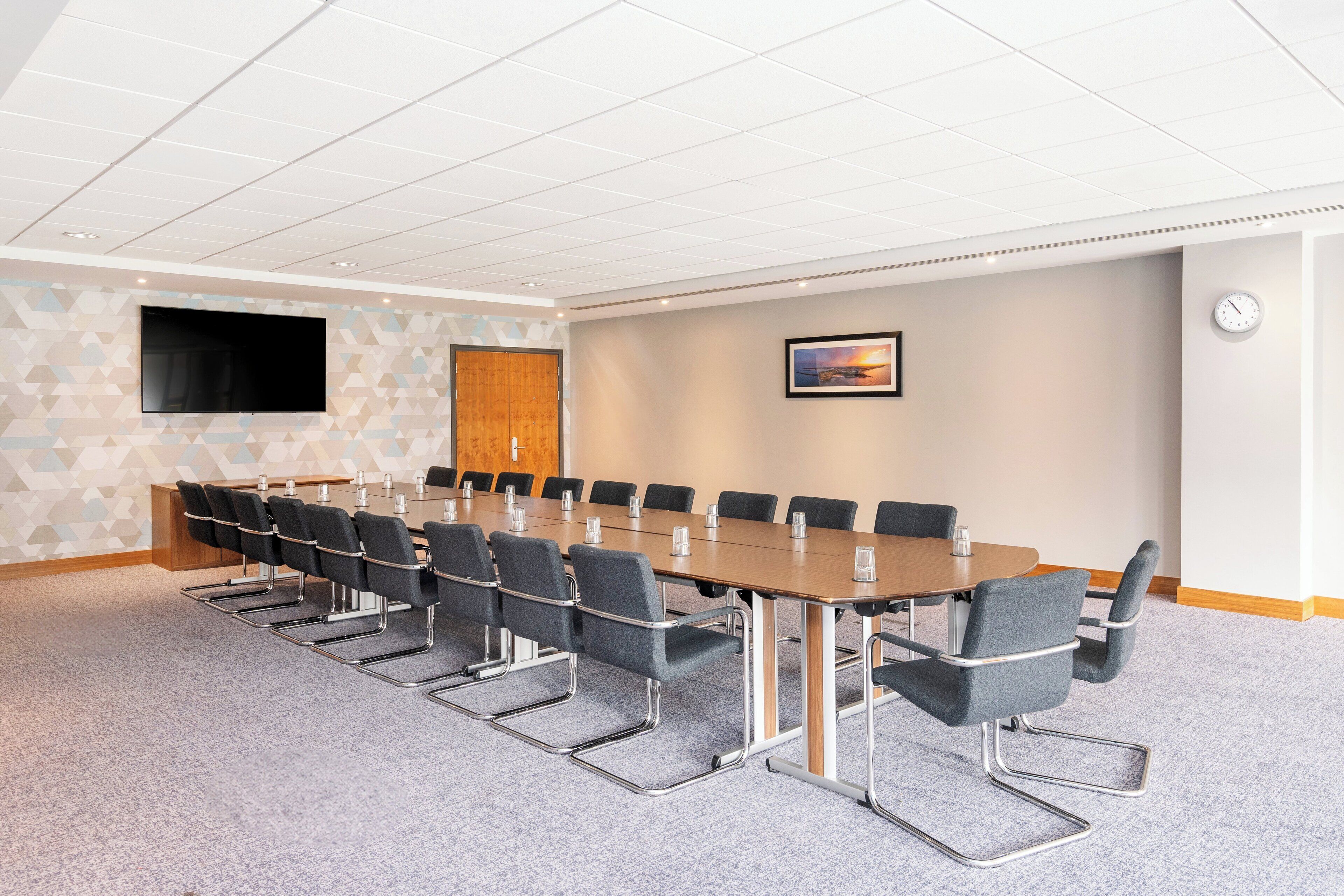 A conference room with a long table, chairs, a flat-screen TV, a clock, and a framed picture on the wall.