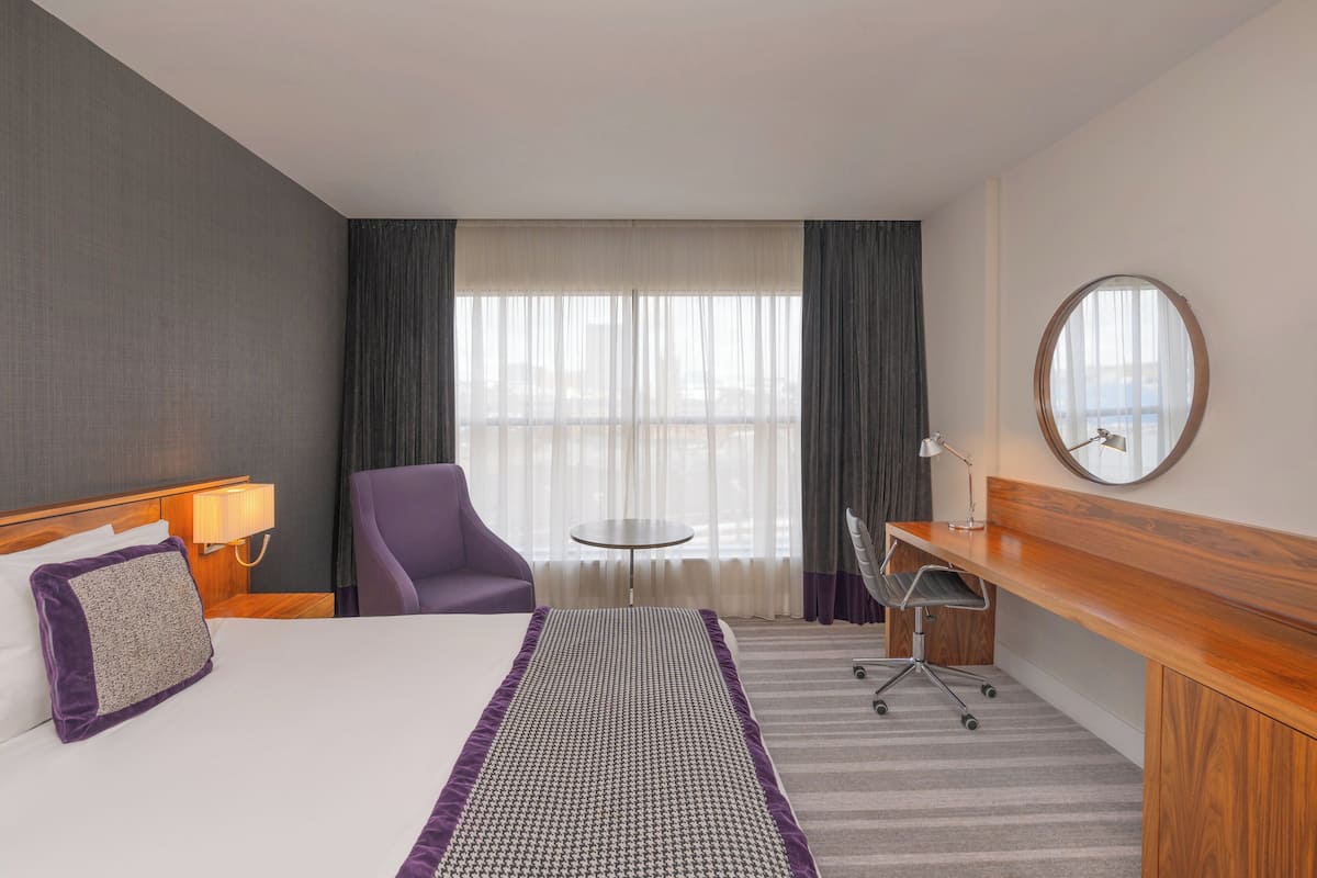 A hotel room with a bed, a desk, a chair, a round mirror, and a view of the city through large windows.