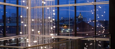 A modern interior with a glass railing and hanging lights, offering a view of the cityscape outside.