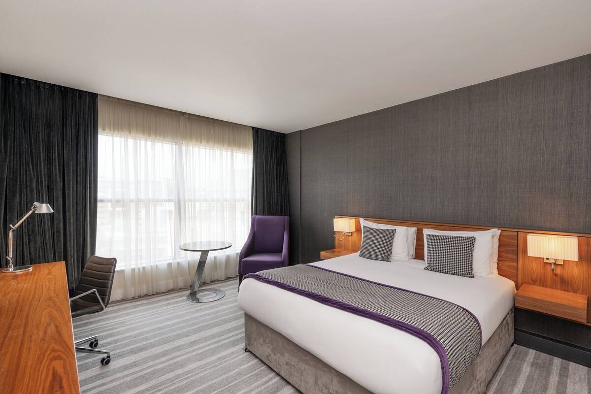 A hotel room with a large bed, a desk with a lamp, a purple armchair, a small round table, and a view of the outdoors through sheer curtains.
