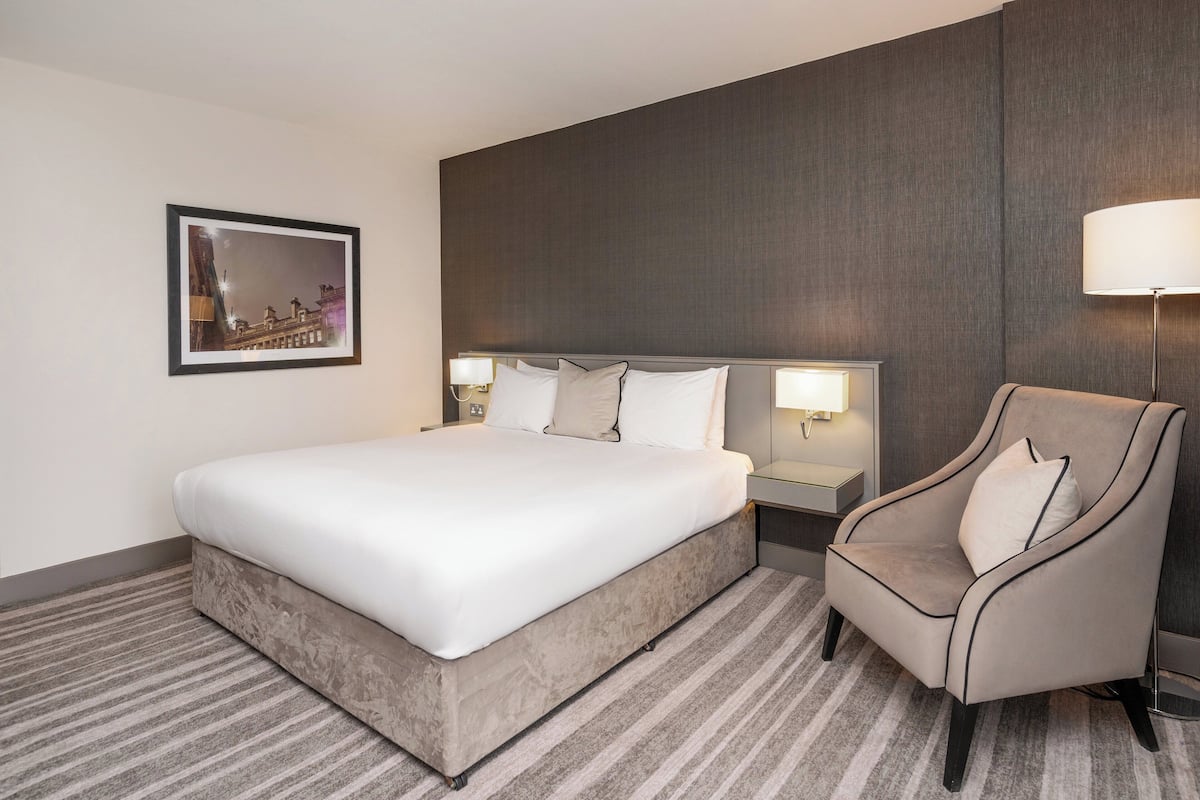 A hotel room with a large bed, a chair, a nightstand with a lamp, and a framed picture on the wall.
