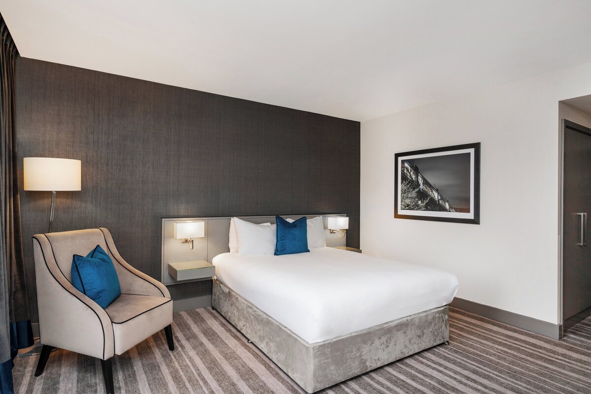 Hotelkamer met een groot bed, een stoel, een nachtkastje, een lamp en een ingelijste foto aan de muur.