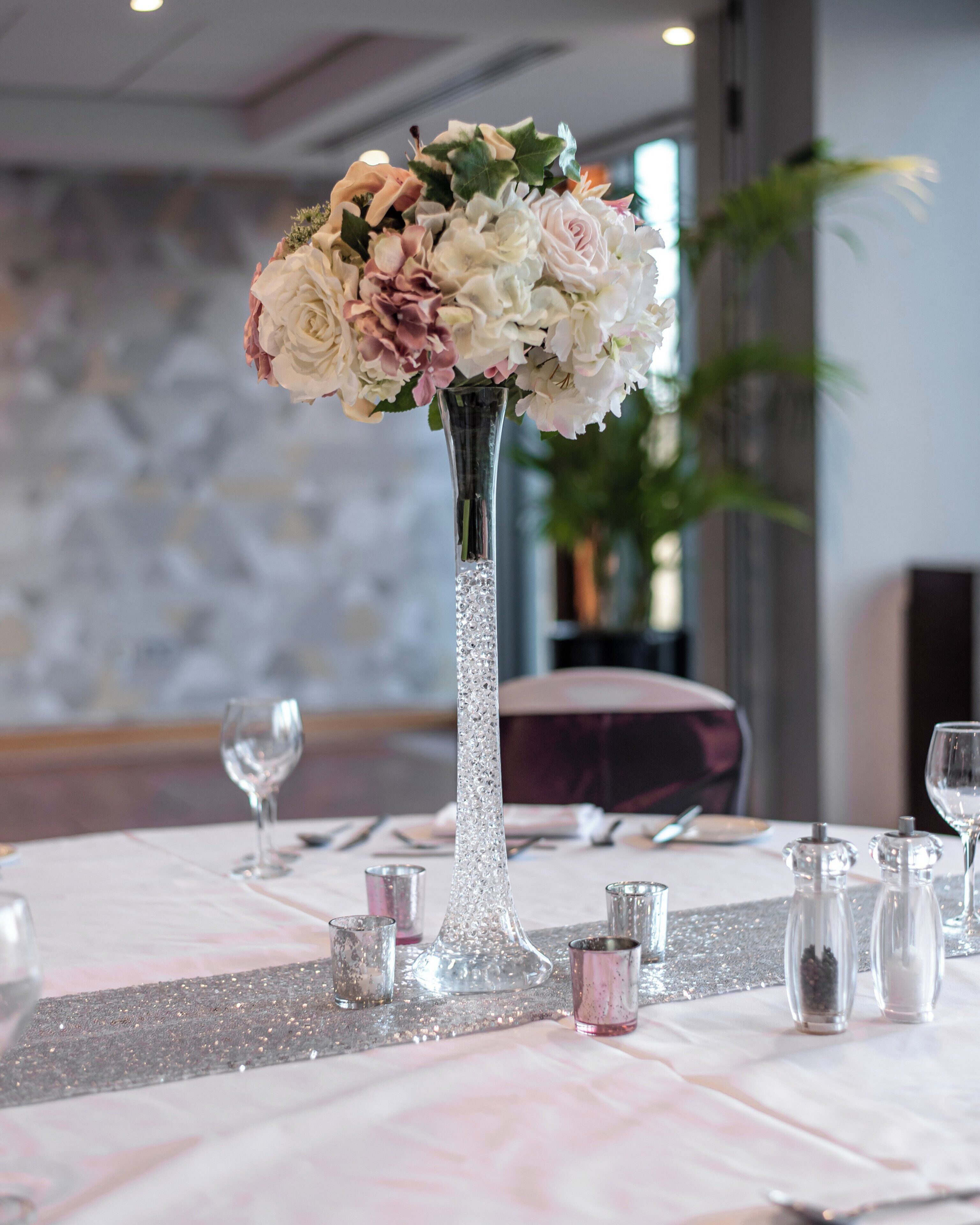 A table set with a clear glass vase containing flowers, small glassware, and a decorative silver fringe.
