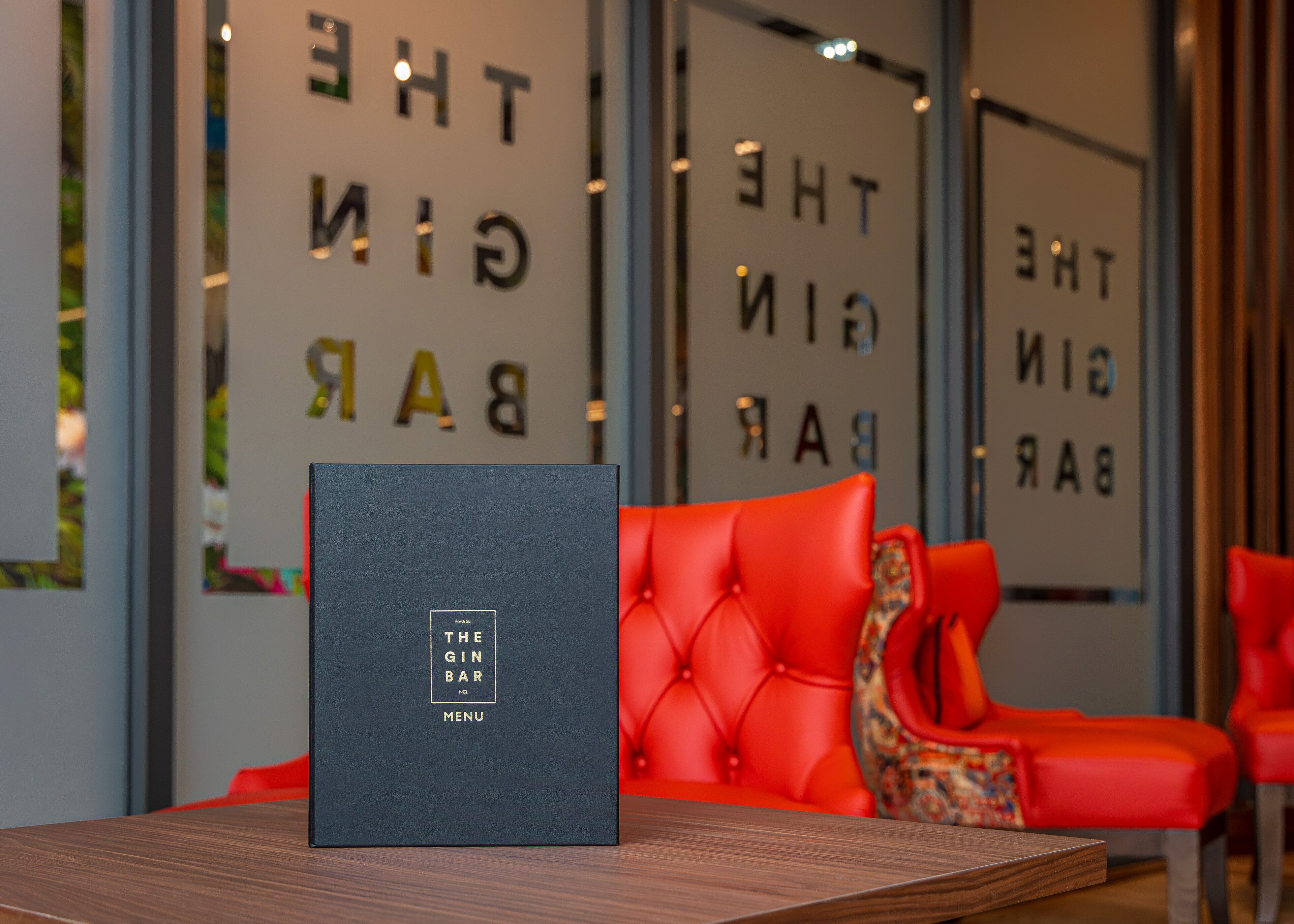 A dark blue menu stand with "The Gin Bar" text in a gold box, placed on a wooden table in front of a red upholstered chair.