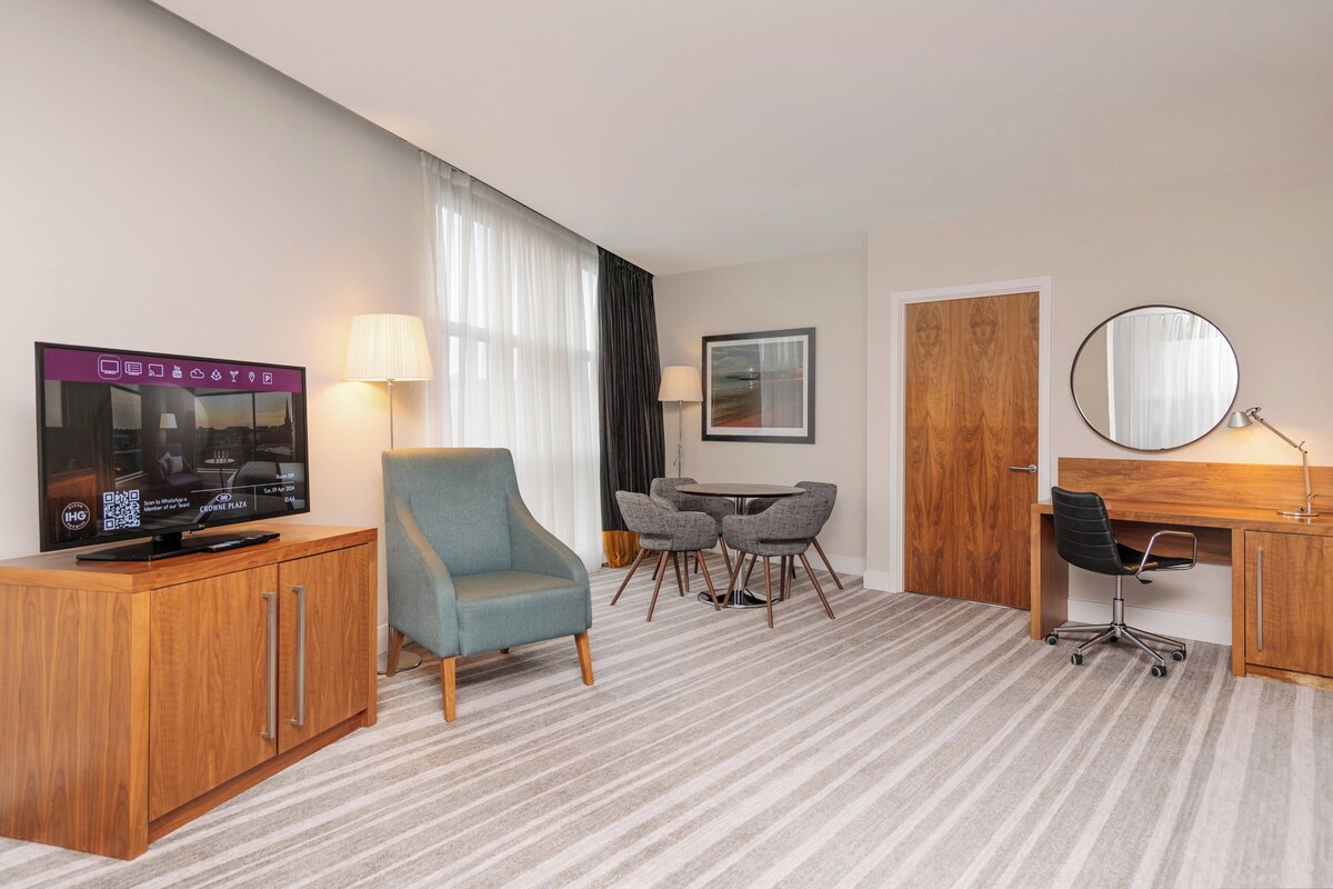 Een hotelkamer met een houten kast, een televisie, een blauwe fauteuil, een eettafel met stoelen, een bureau met stoel, en een ronde spiegel.