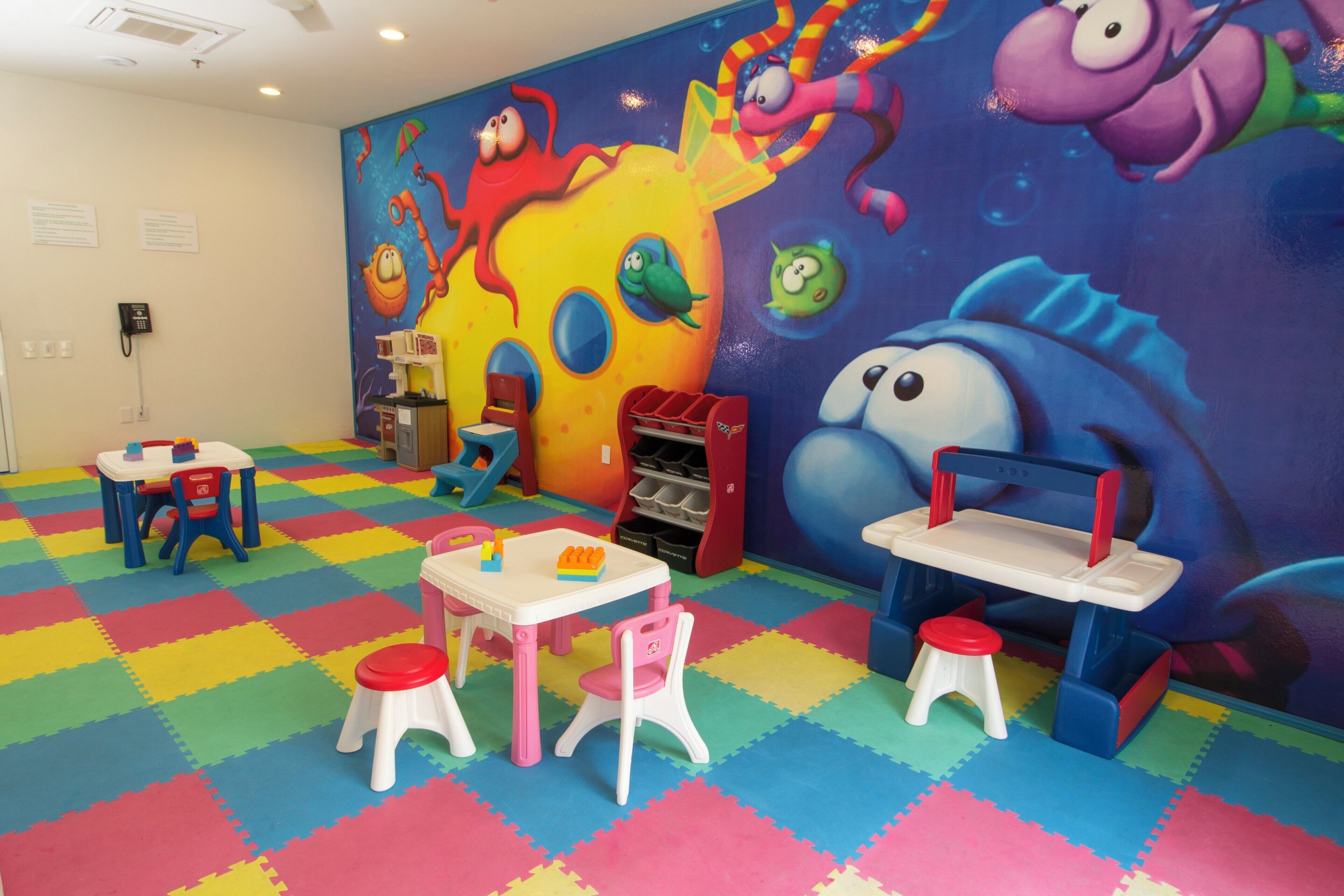 Un salón infantil con coloridos tapetes de espuma, mesas de juego, sillas y un mural llamativo en la pared.